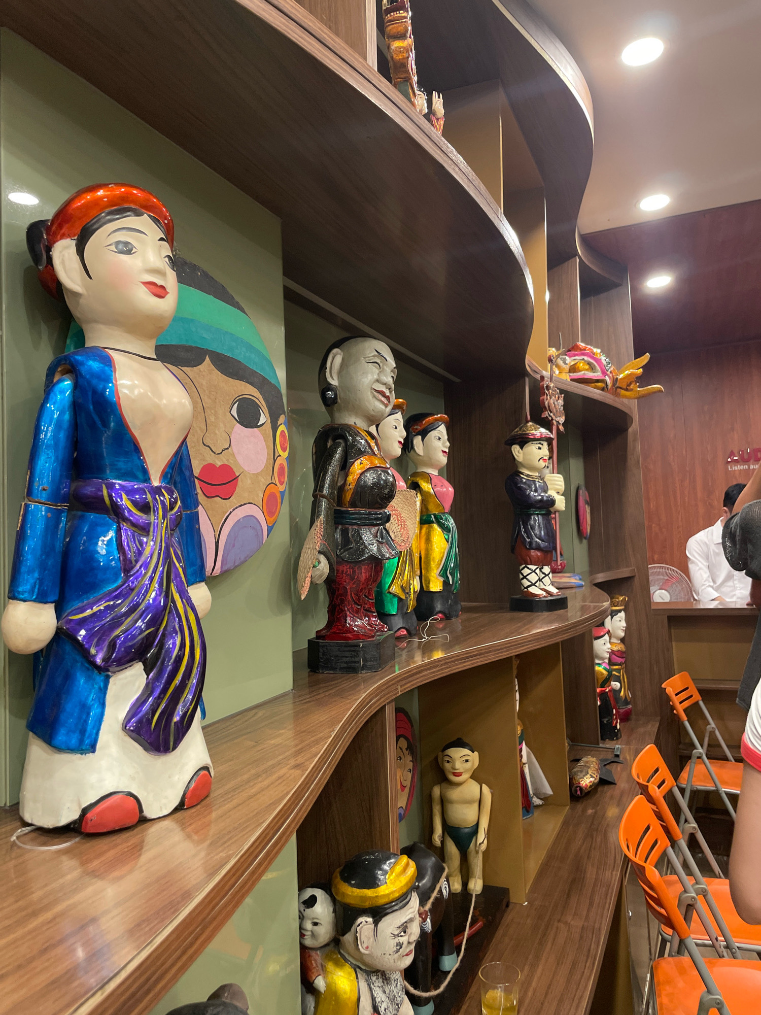 This image shows a display of Vietnamese water puppets in what appears to be a shop or museum in Hanoi, Vietnam. The puppets are displayed on wooden shelves. In the foreground, a large female puppet in a blue and purple robe stands prominently. To its right, several smaller puppets are arranged, including one with a black robe and red pants, and another in a yellow and green outfit. Further down the shelves, more puppets are visible, some in traditional Vietnamese attire. In the background, a person in a white shirt can be seen near a desk, and orange chairs are stacked to the right. The overall environment suggests a commercial or cultural space dedicated to showcasing these traditional art forms.
