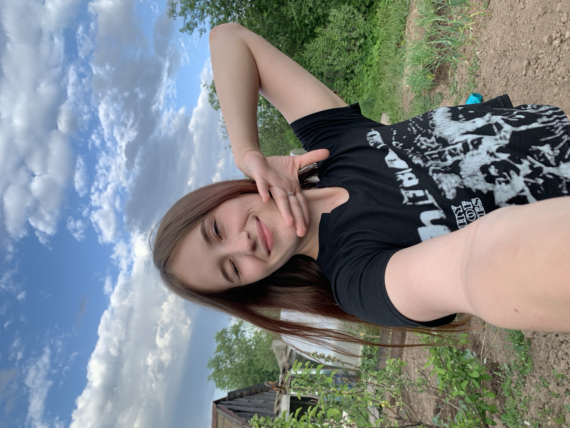 A young woman poses for a selfie outdoors in Фоки, Russia. She is wearing a black t-shirt with a white graphic and a ring on her finger. The woman is holding her arm up, with her hand near her face. In the background, there is a sky filled with blue and white clouds. Green foliage and an old wooden structure are also visible.