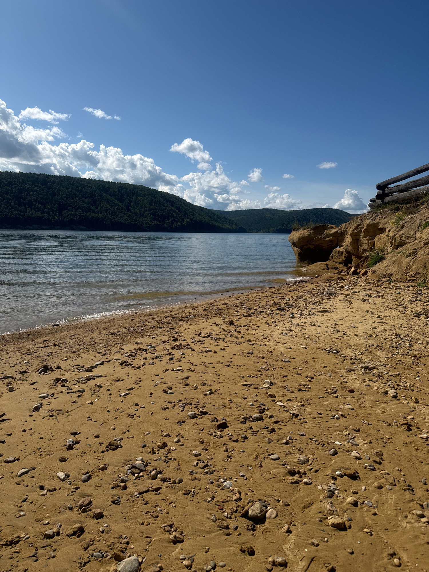 The image depicts a sandy beach with scattered pebbles leading to a calm body of water. In the background, forested hills rise under a blue sky with scattered white clouds. A wooden fence is visible on the right side of the beach, overlooking the water and the distant hills. The scene is natural and serene, characteristic of a lakeside or riverside location. The specific location in the city of Ангара (база Отдыха), Russia, is not visually identifiable from the image itself without further context or landmarks.