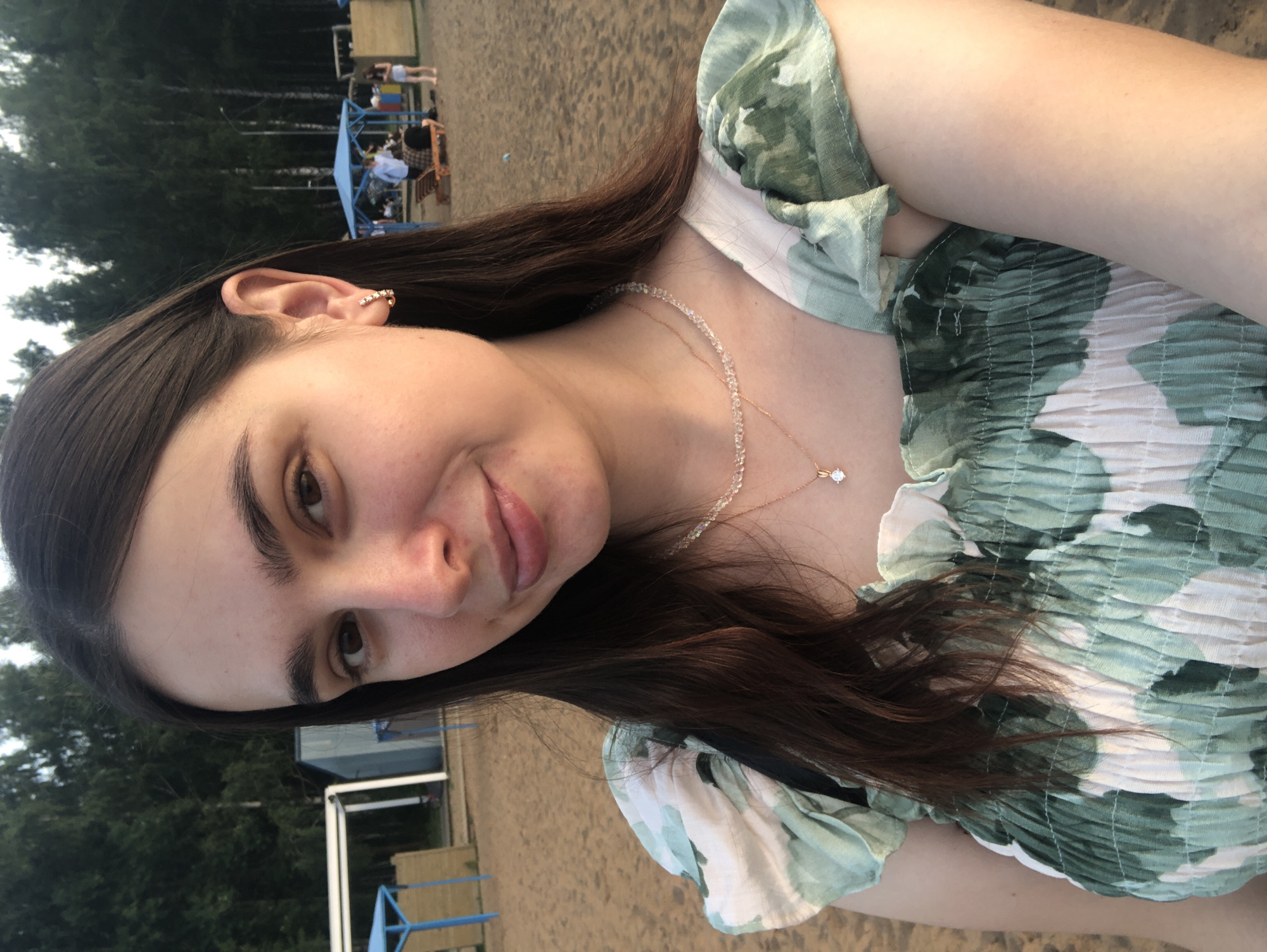 A young woman with long, dark hair and brown eyes is taking a selfie on a sandy beach. She is wearing a green and white floral print dress with ruffled sleeves. She has a delicate necklace with a pendant and small hoop earrings. In the background, there are trees, a blue canopy structure, and other people on the beach. The scene suggests a recreational setting, possibly a park or public beach area. The lighting indicates it is likely late afternoon or early evening. The location is the bank of the Kama River in Chaikovsky, Russia.