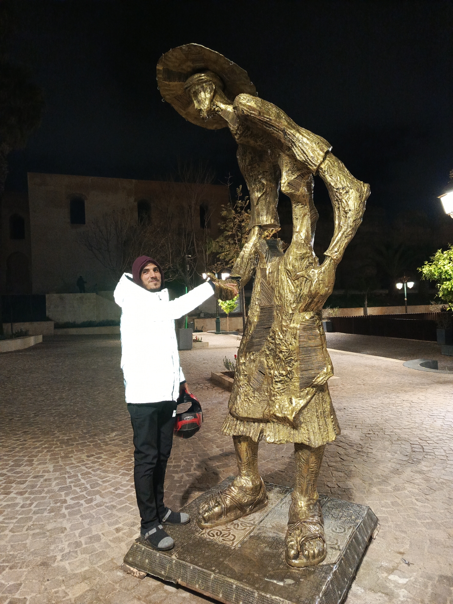 A man in a white jacket, black pants, gray shoes, and a red beanie stands next to a large, golden statue on a cobblestone surface. The man holds a helmet in his left hand and extends his right hand towards the statue. The statue depicts a figure with a wide-brimmed hat and what appears to be a tunic. In the background, there is a building, trees, and streetlights. The scene is at night. The location is Al Machouar - Stinia, Morocco.