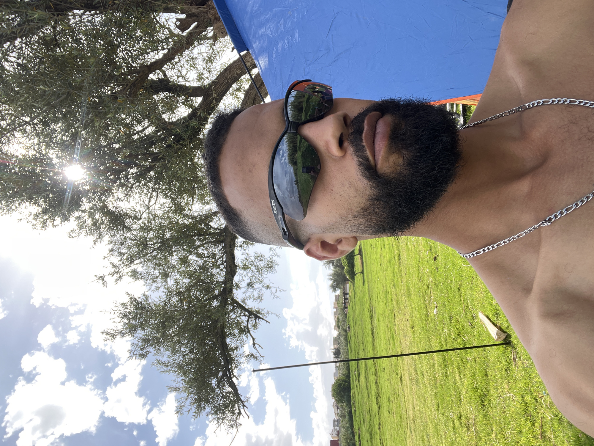 A close-up, low-angle shot shows a light-skinned man wearing sunglasses and a silver chain, with a beard. He is situated outdoors, as indicated by the blue sky and puffy white clouds visible in the background. The reflection in his sunglasses suggests a grassy field and trees. A tree with leafy branches is visible to the left of the frame, and the sun shines through its leaves. A piece of white debris is seen on the green grass in the foreground. Based on the reflections and landscape, this photograph may have been taken in a recreational area or park in Dkhissa, Morocco, where green spaces are found within the city.