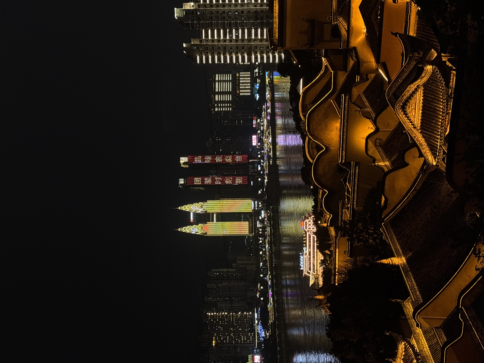 At night, Yuzhong District, China, is illuminated with lights reflected on the river. The buildings and skyline glow, creating a vibrant urban scene. Traditional Chinese architecture contrasts with modern skyscrapers. A boat, also illuminated, adds to the dynamic scene.