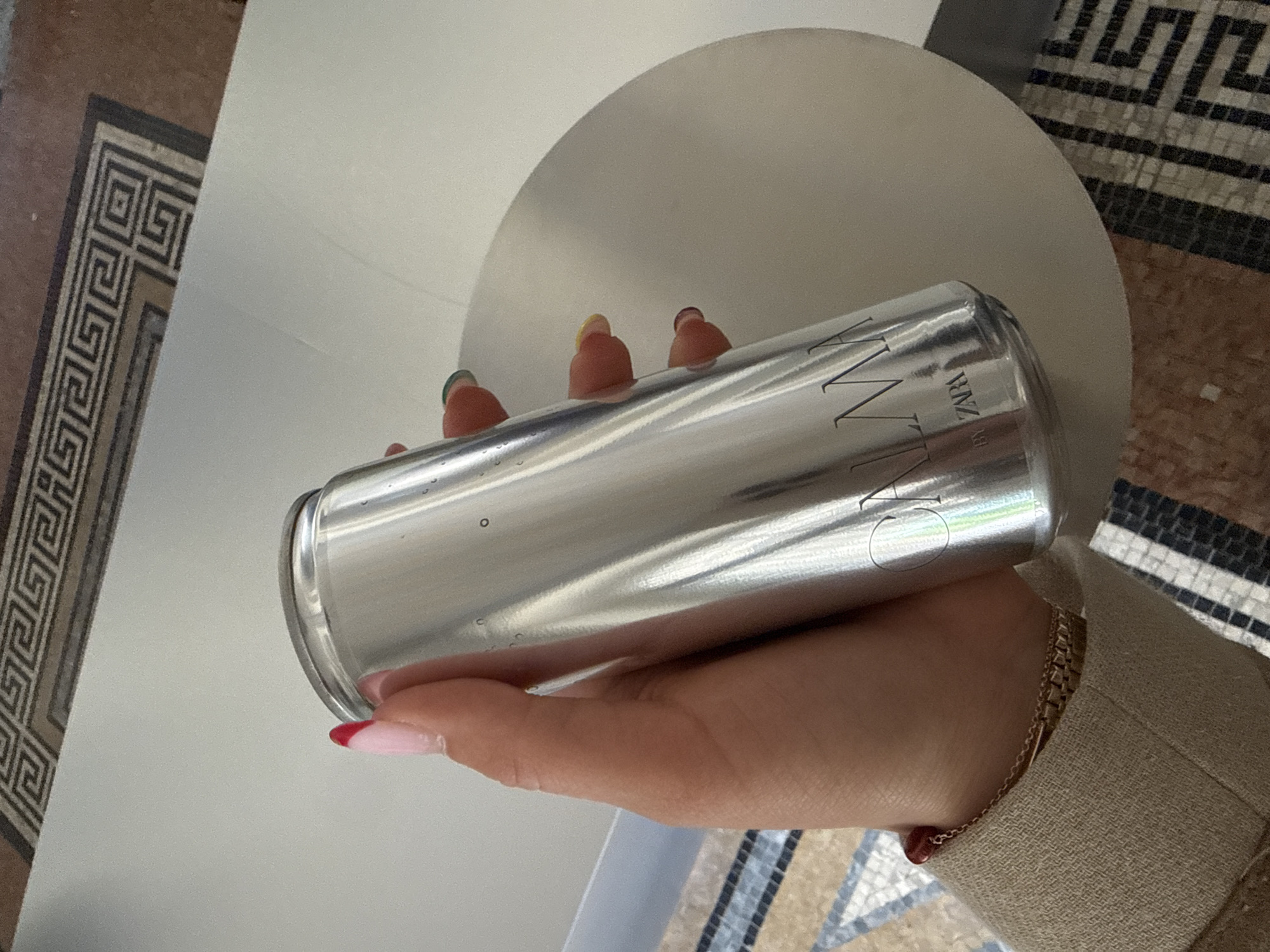 The image displays a close-up, slightly tilted view of a person's hand holding a sleek, reflective silver can. The hand has manicured nails, with some fingernail tips painted red and others green. A delicate gold-colored chain bracelet adorns the wrist, which is partially obscured by the cuff of a light brown or beige sleeve, likely from a jacket or blazer.
The silver can features prominent text, with the word "OWW" clearly visible in large, stylized capital letters. Beneath this, in a smaller font, the text "BY ZARA" is discernible, identifying the brand of the product. The can's metallic surface reflects light and shows subtle textures and possible small droplets.
The background consists of two main elements. To the right and upper portion of the frame, a large, light-colored, possibly circular or curved surface is present. To the left and bottom right, an intricate tiled floor is visible. This floor showcases a distinctive geometric pattern, strongly resembling a Greek key or meander design, rendered in dark (black), light (white), and brown-toned tiles. This architectural detail is consistent with the provided context of the location being Milan, Italy, suggesting an indoor setting with classic design elements. The image captures a moment of interaction with the branded can against an aesthetically rich backdrop.