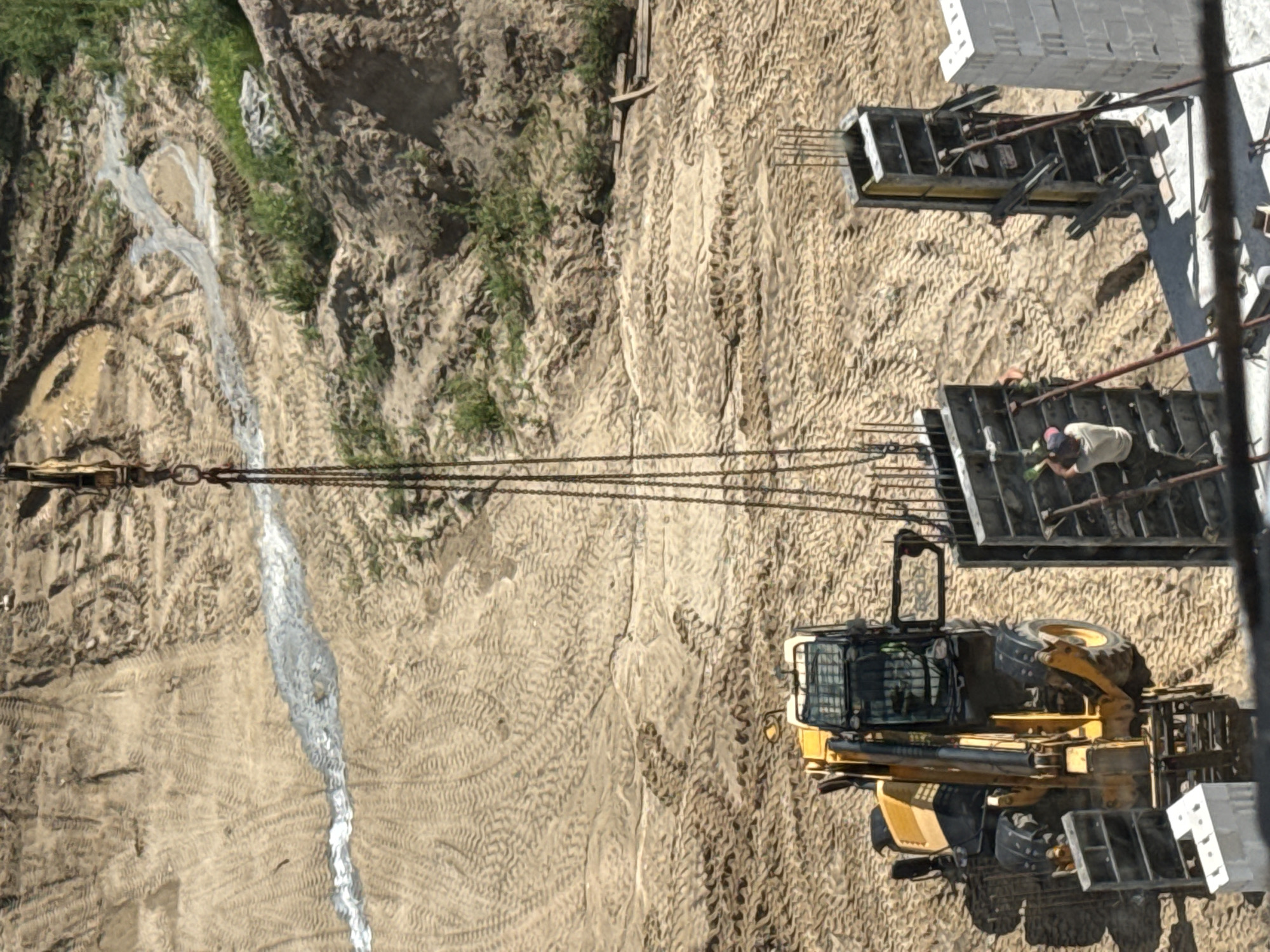 This aerial, top-down view captures a construction site in Mikołajki, Poland. The scene is dominated by a sandy, uneven terrain with numerous tire tracks indicating heavy machinery activity. A bright yellow excavator is parked on the sand, its boom arm extended. In the background, several large concrete formworks, reinforced with rebar, are visible. One worker, wearing a light-colored shirt and a cap, is actively engaged with the formwork, possibly preparing it for concrete pouring. The lighting suggests it's daytime, with distinct shadows cast by the machinery and structures. A thin, white stream, possibly water or a cement mixture, flows down a slight incline on the left side of the frame, cutting through the sandy ground. The overall impression is one of ongoing construction work under clear daylight conditions. No visible text is present in the image.