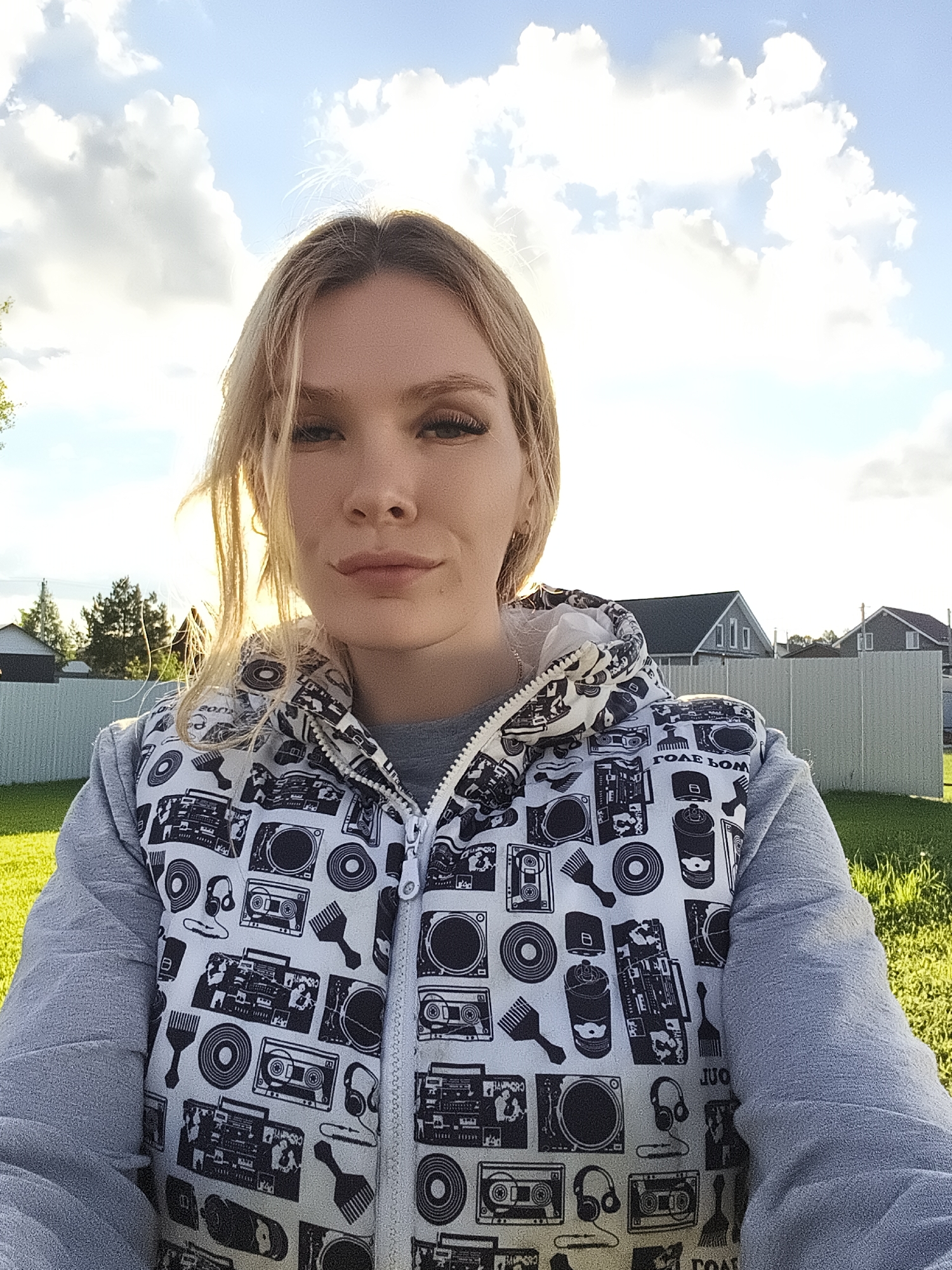 A woman with light blonde hair and fair skin is the primary subject of a selfie-style photograph taken outdoors in what appears to be a residential area. The woman is wearing a gray, long-sleeved sweatshirt under a zip-up vest featuring a white background with black designs of boomboxes, records, and other urban-themed images. The sky is visible in the background, showing patches of blue interspersed with white clouds. Beyond the woman and the grassy area, there are houses with dark gray or black roofs and light-colored siding, separated by a light-colored fence. The scene is likely in the city of Русский Вожой, Russia, based on the prompt's instructions.