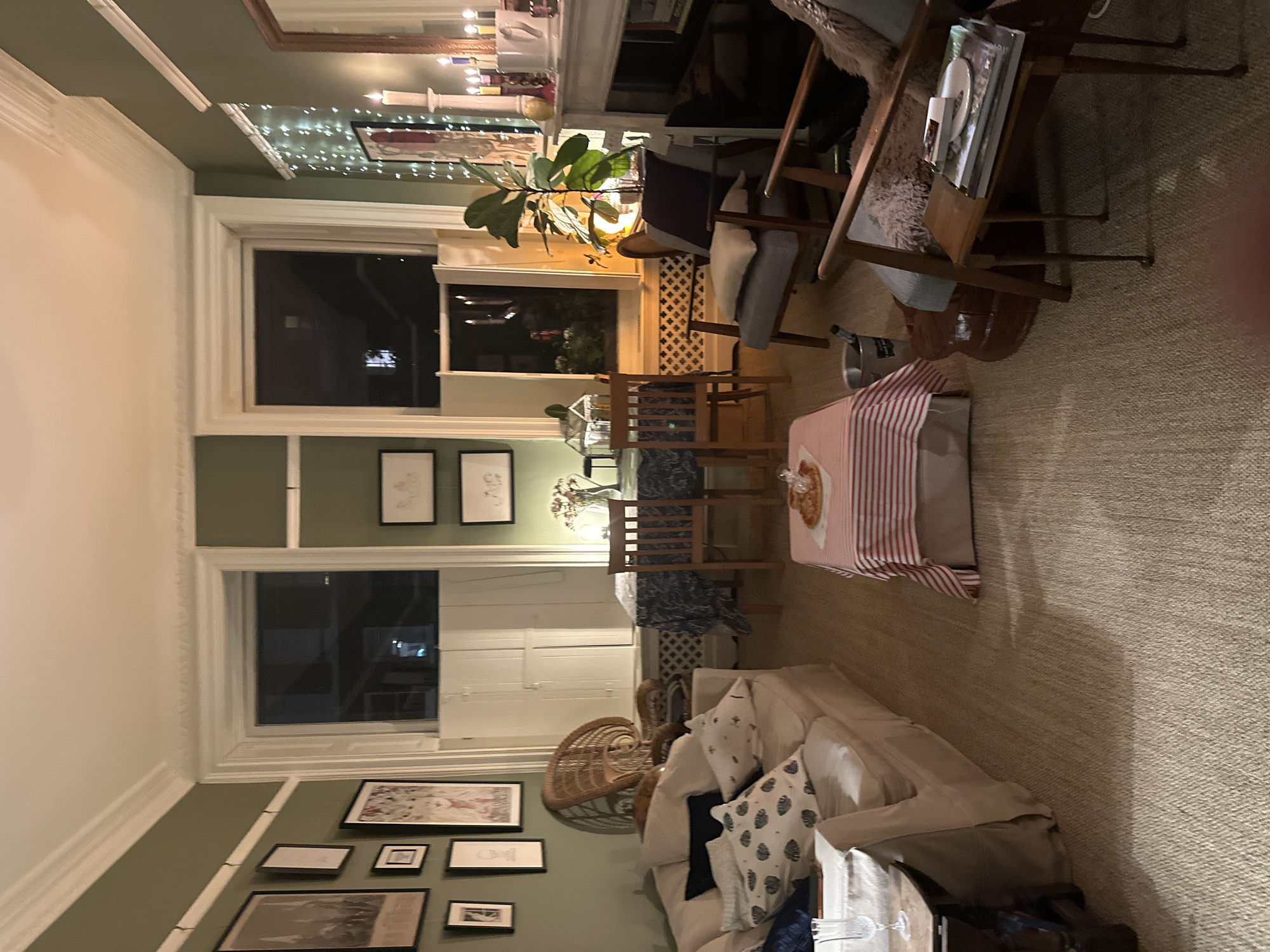 The image depicts an interior view of a living room in a domestic setting. Natural light enters through two large windows, suggesting it is either dawn or dusk. The room is decorated with an eclectic mix of furniture, including a light-colored sofa with patterned cushions, wooden chairs, and a table covered with a red and white striped tablecloth. Various framed artworks adorn the walls. A plant sits near one of the windows. The flooring is a neutral-colored carpet. No people are visible in the image. The location is likely in the city of London, United Kingdom, suggested by the architectural style and decor.