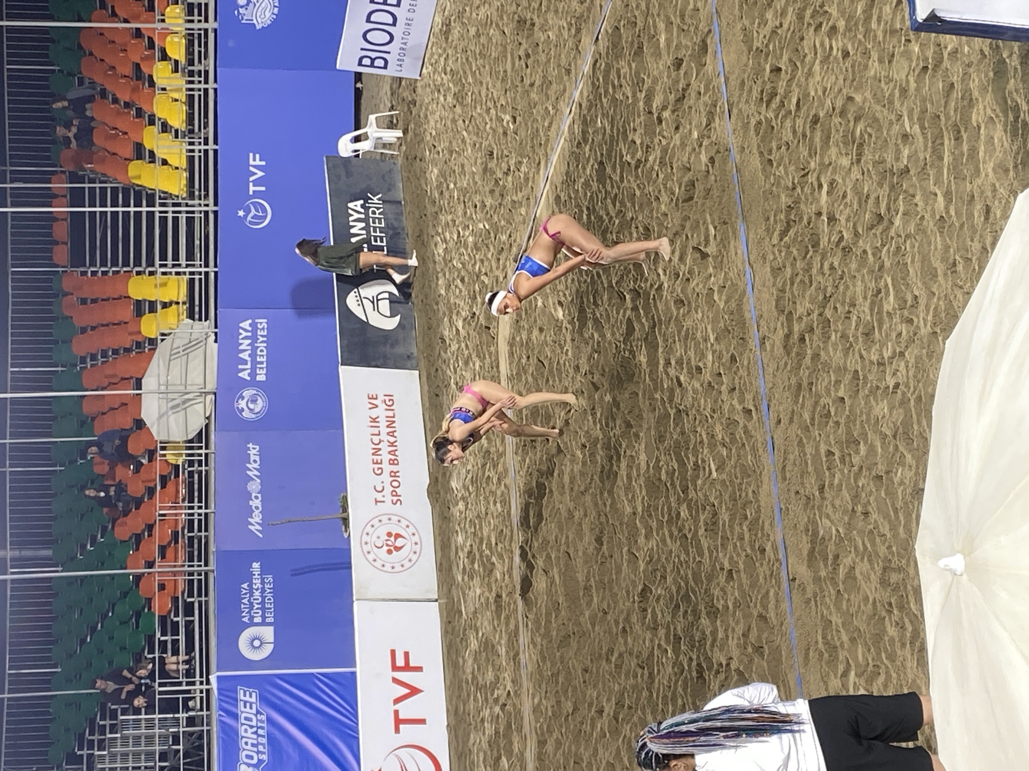 The image captures a beach volleyball scene, likely in Alanya, Turkey, based on the signage. Two female athletes are actively playing on a sandy court, positioned near the center of the frame. The backdrop includes stadium seating, mostly empty, and advertising panels with logos such as "TVF," "MediaMarkt," and "T.C. Gençlik ve Spor Bakanlığı" (Turkish Ministry of Youth and Sports). In the foreground, parts of a white parasol and a person with dreadlocks are visible, suggesting spectatorship. The lighting indicates either late afternoon or early morning.