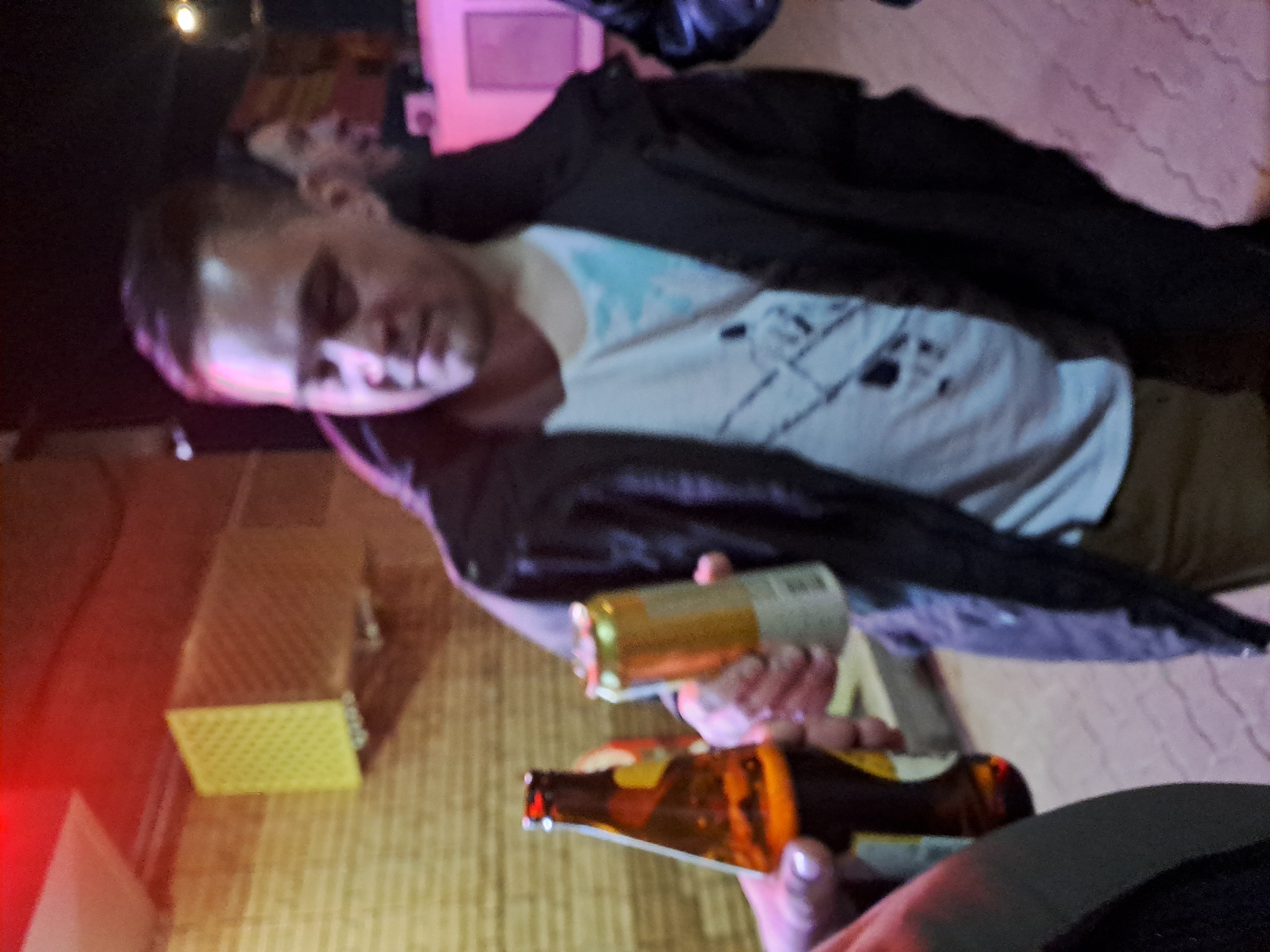 The image depicts a low-light scene in Minino, Russia, featuring three identifiable individuals interacting with beverages. A central male figure, with short, dark hair, wearing a dark jacket over a white t-shirt with a faint pattern, and light brown trousers, holds a gold and silver beverage can extended in his right hand. To his immediate left, partially visible in the lower foreground, another person's hand holds a brown glass bottle with a red-orange label, positioned as if in interaction with the central figure's can. A blurry male face, visible in profile, is present behind the central figure's right shoulder, looking to the right. A fourth individual's dark-clad shoulder is partially visible on the far right. The background consists of a light brown, textured wall. Mounted on this wall in the upper left is a yellow, rectangular object with a grid pattern, possibly a ventilation cover or acoustic panel. The environment is dimly lit by ambient red and pink light sources, with a distinct red light emanating from the upper left and a bright pink light casting a glow on a rectangular object or surface in the far right background. The ground in the foreground appears to be a reddish-pink tiled or paved surface.