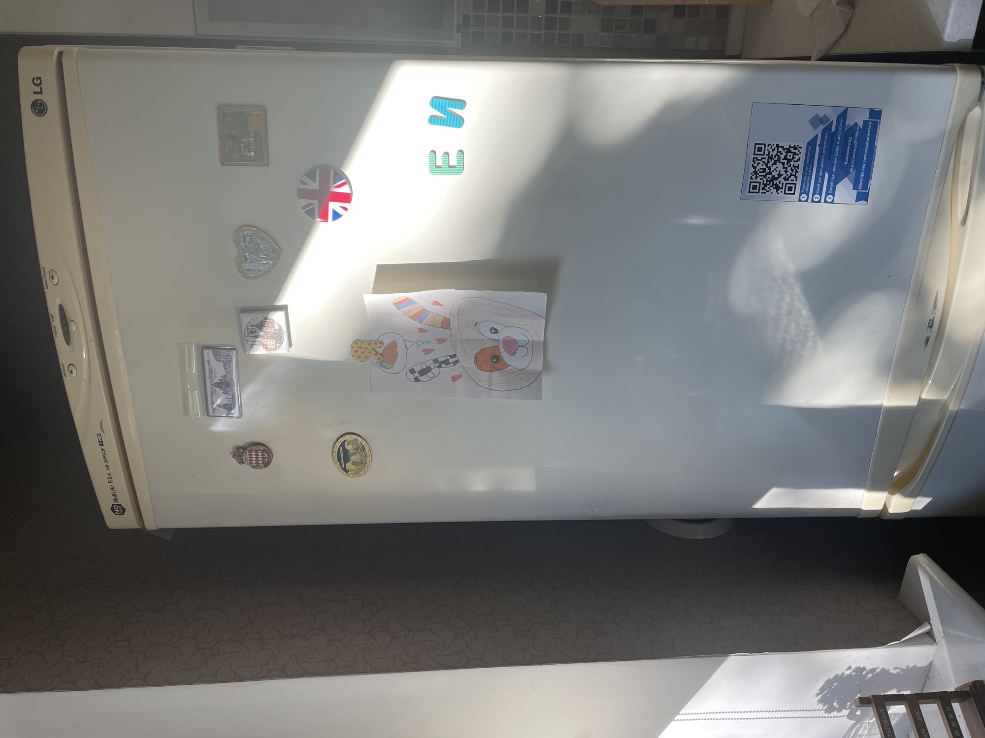 The image shows a white LG refrigerator in a domestic setting. Several magnets and a child's drawing are attached to its surface. There are two magnetic letters, "E" and "N", on the right side of the refrigerator door. A QR code sticker with Russian text is affixed to the lower right portion of the door, indicating it pertains to Kaliningrad, Russia and providing a URL for "concierge39.v1da.ws". The refrigerator is positioned in a well-lit room, with sunlight casting shadows across its surface. A dark patterned wallpaper is visible on the wall behind the refrigerator.