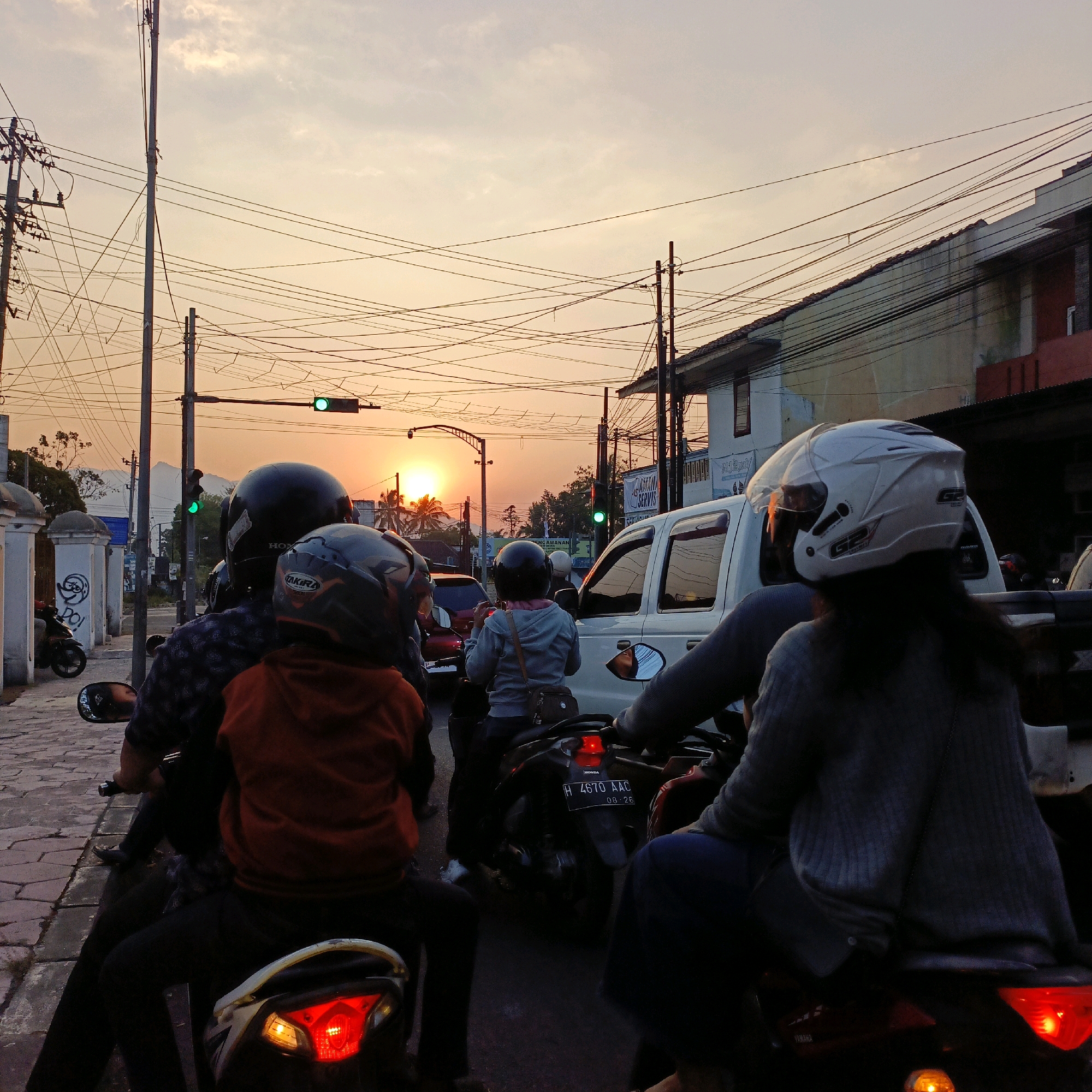 This image depicts a traffic scene at dusk in Salatiga, Indonesia, near a traffic light. Multiple motorcycles, cars, and a white van are visible. The sky is a warm orange, indicating the setting sun, which is partially obscured by palm trees and buildings. Power lines crisscross the sky, a common sight in urban Indonesian landscapes.
In the foreground, several individuals are riding motorcycles. One motorcycle has two people, the front rider wearing a black helmet and the rear passenger wearing an orange hooded jacket. Another motorcycle in front of them has a rider wearing a blue hooded jacket. Further ahead, people on other motorcycles and in cars are waiting, likely at the traffic light.
The environment appears to be an urban street with sidewalks, buildings lining the road, and streetlights. The overall impression is a busy, everyday commute during sunset in a Southeast Asian city.