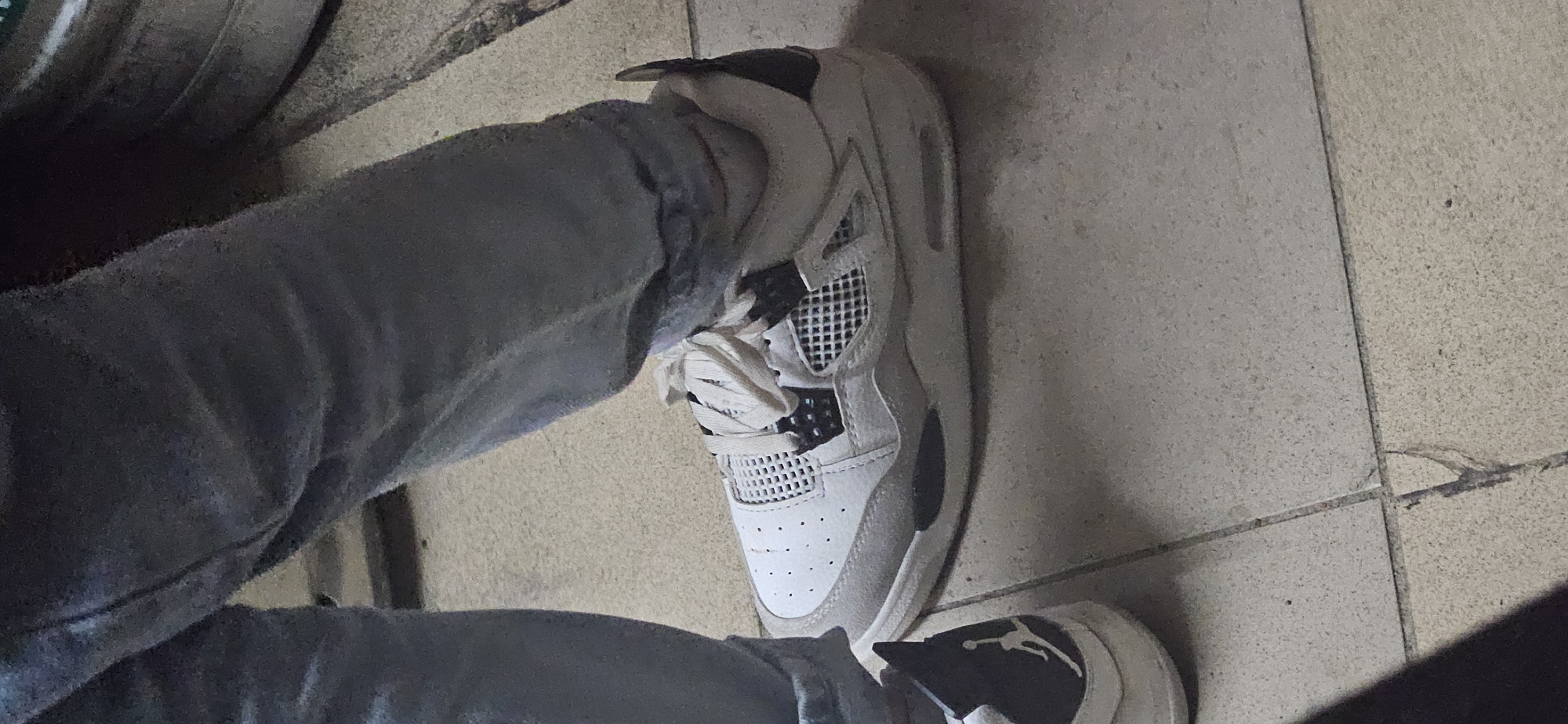 The image depicts a person wearing grey jeans and two different Air Jordan 4 sneakers. The right shoe is predominantly white with black accents, while the left shoe is not fully visible, but the heel appears to be black with a white Jumpman logo. The person is standing on a tiled floor. The location cannot be determined from the image.