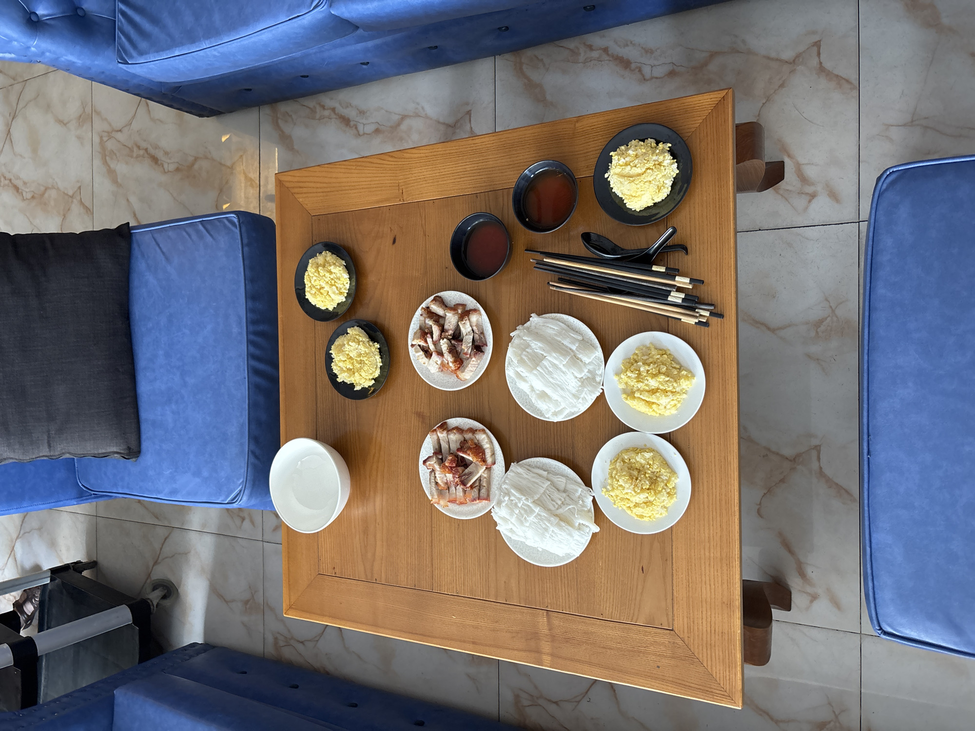 The image captures a close-up, overhead view of a wooden coffee table laden with food, set within a brightly lit indoor space. The table features a collection of small plates and bowls containing what appears to be a meal. Specific items include sliced roasted meat, a white, shredded food item, small portions of a yellow, granular substance, and two dark bowls filled with a reddish liquid, likely a sauce or dipping liquid. A set of chopsticks and a black spoon are also present.
The table itself is rectangular with a light wood finish and has carved legs. It is situated on a tiled floor with a marble-like pattern in shades of beige and brown. Surrounding the table are sections of blue upholstered furniture, one of which has a dark gray cushion. In the background, a portion of a black rolling cart with metallic legs and wheels is visible, suggesting an indoor setting such as a restaurant, cafe, or residential dining area. The lighting appears to be natural, possibly from a nearby window, casting subtle shadows. The overall impression is one of a prepared meal, ready to be served or consumed. The presence of specific cuisine elements suggests a Vietnamese setting, potentially in Nha Trang Ward, Vietnam.