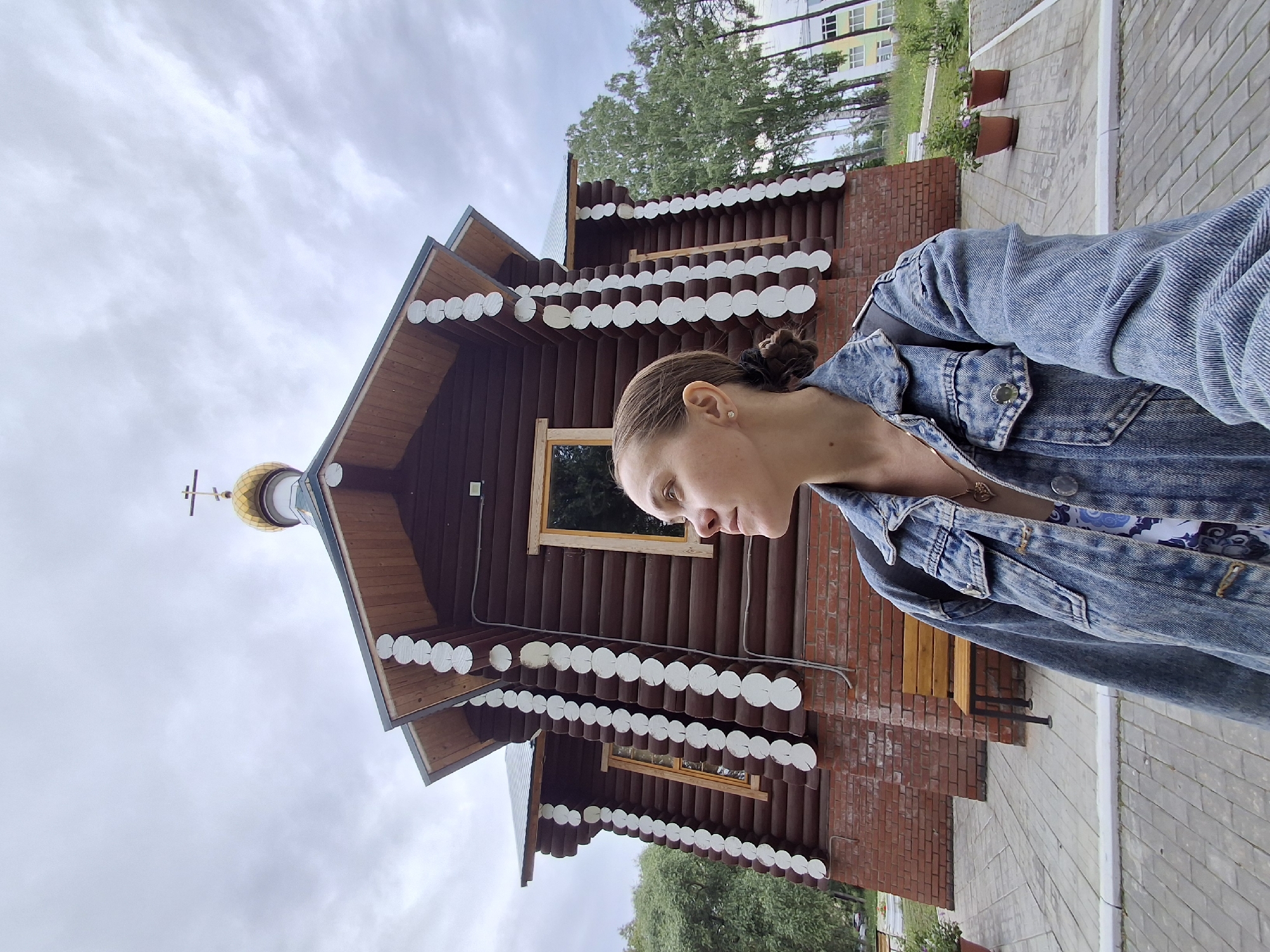 A young woman with her hair pulled back in a bun is standing in front of a wooden church in Chaikovsky, Russia. She is wearing a denim jacket over a patterned top. The church is made of dark brown logs with white decorative trim around the roofline. It has a golden dome with a cross on top. The building is constructed on a brick foundation. To the right of the church, a paved walkway leads to a modern building in the background. Two large potted plants are visible near the walkway. The sky is overcast, suggesting a cloudy day. The image appears to be a selfie, with the woman looking down and to the side. The time of day is difficult to determine due to the lighting conditions.