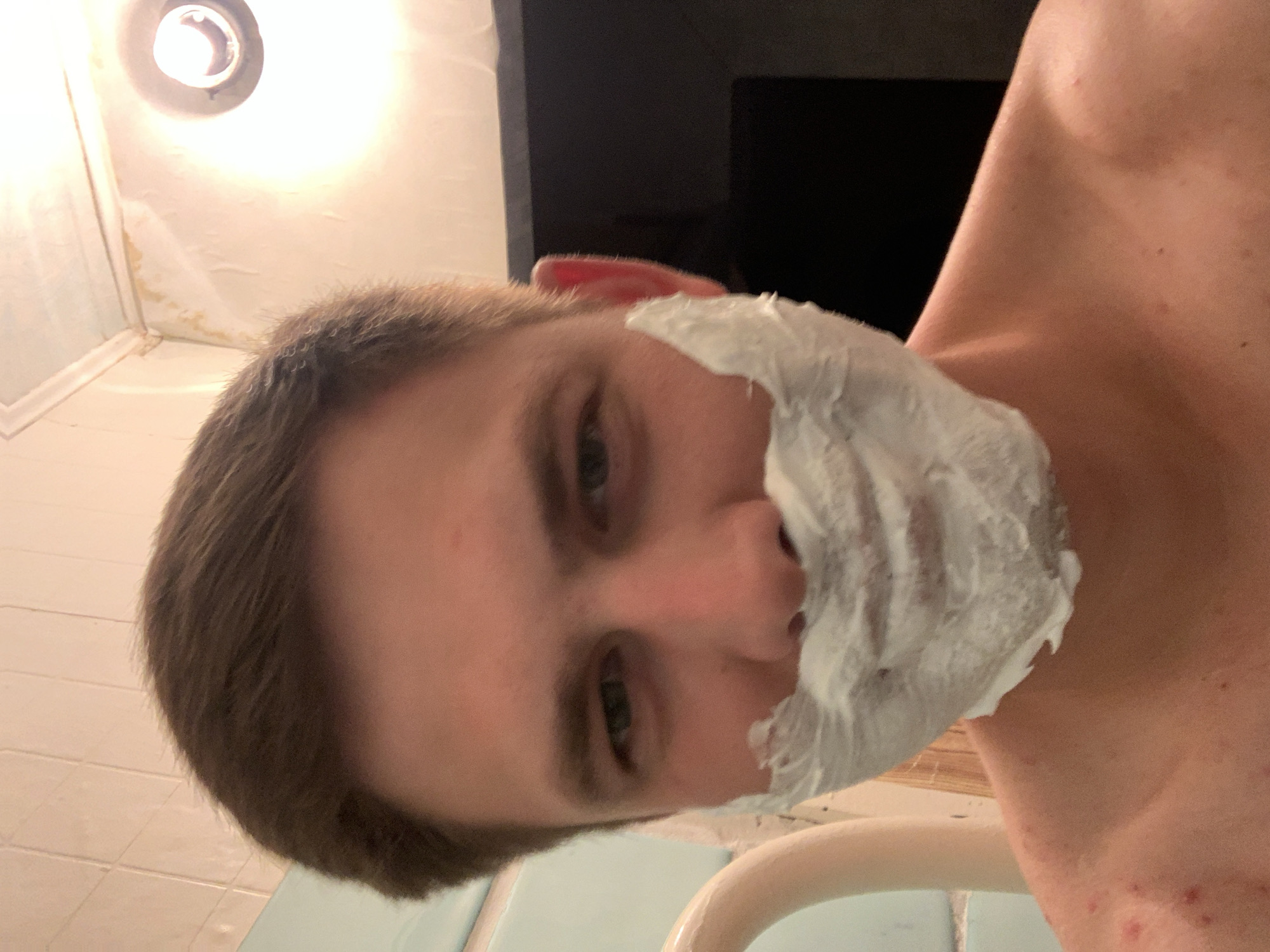 A close-up, selfie-style shot shows a person with short brown hair and a face covered in shaving cream. The person's eyes are open, and they are looking directly at the camera. The background consists of a tiled wall and a light fixture. There is a curved object, possibly a pipe or handle, visible on the left side of the frame. The lower part of the image shows the person's bare shoulders and upper chest, with some visible red marks. The scene appears to be in a bathroom, likely a shower or tub area, given the tiles and lighting. The image is grainy and appears to have been taken in low light conditions.
