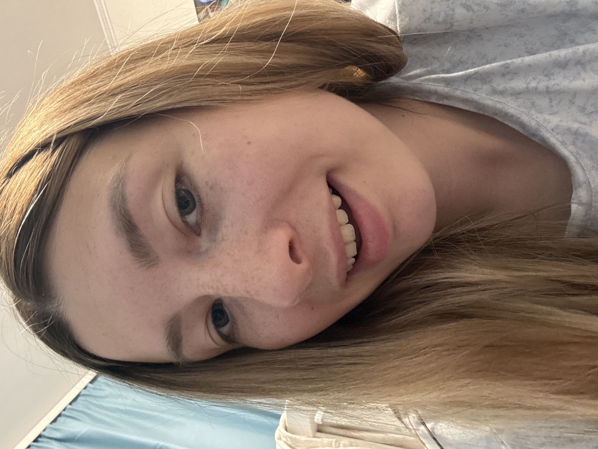 A close-up, slightly angled portrait shows a young woman with fair skin, freckles, and light brown hair. She has light-colored eyes and is smiling, revealing her upper teeth. She is wearing a light-colored top with a subtle pattern. The background includes a white wall, a blue curtain or fabric, and a glimpse of what appears to be bedding or other soft furnishings. There is no indication of the location being Moscow, Russia.