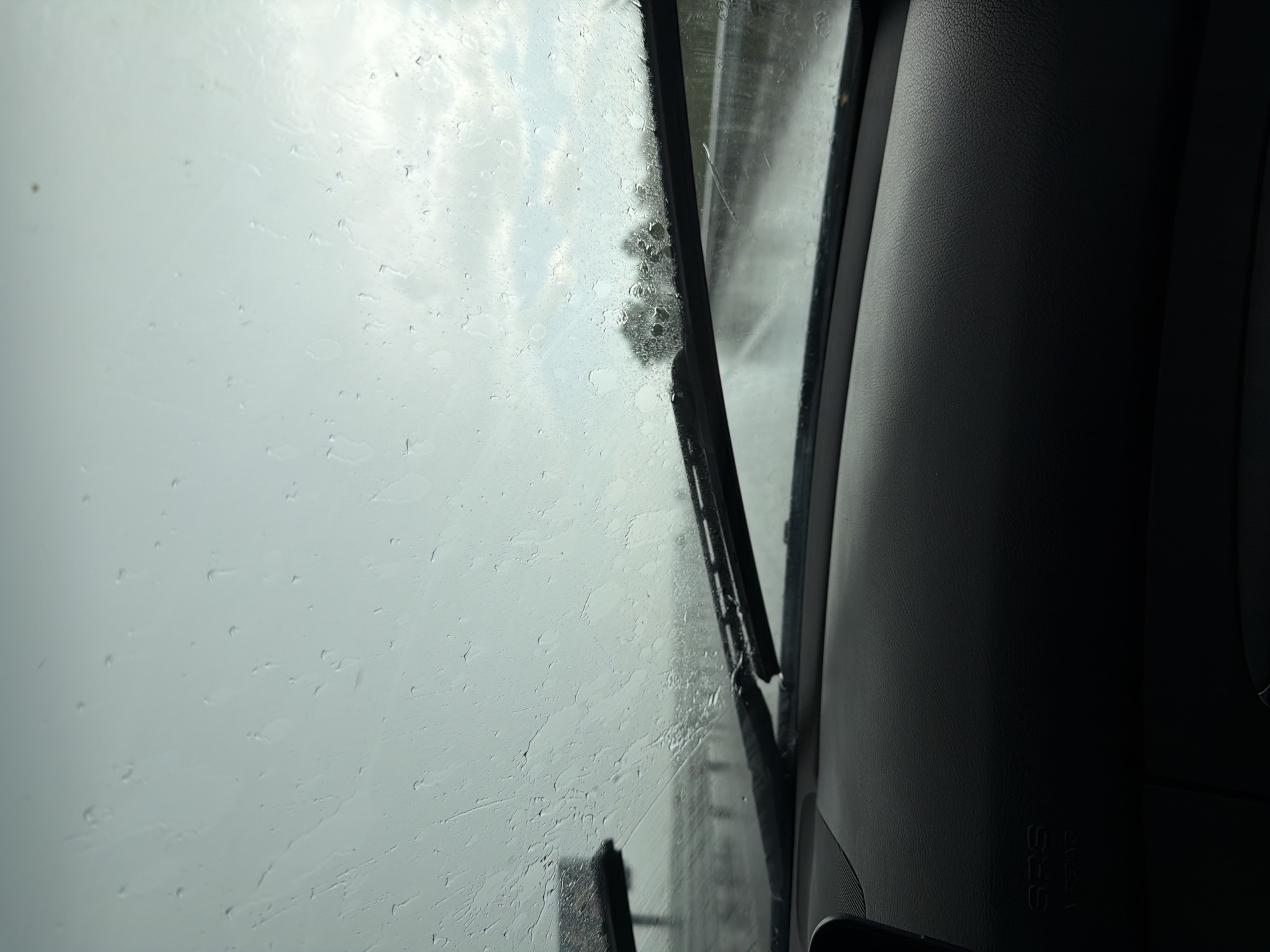 The image shows a view from inside a vehicle, likely a car, through the windshield. The windshield is covered in raindrops, indicating it is raining. A windshield wiper is partially visible, positioned vertically in the center of the frame. Outside, through the blurred and rain-streaked glass, the scene appears overcast and gray. The presence of a tree can be discerned in the distance.
The right side of the image is dominated by the interior of the vehicle, specifically the dashboard, which is dark gray or black and textured. An "SRS AIRBAG" label can be seen on the dashboard. The scene does not provide enough information to establish a location in Malko Tranovo, Bulgaria. No people, specific objects, actions, or interactions are visible.