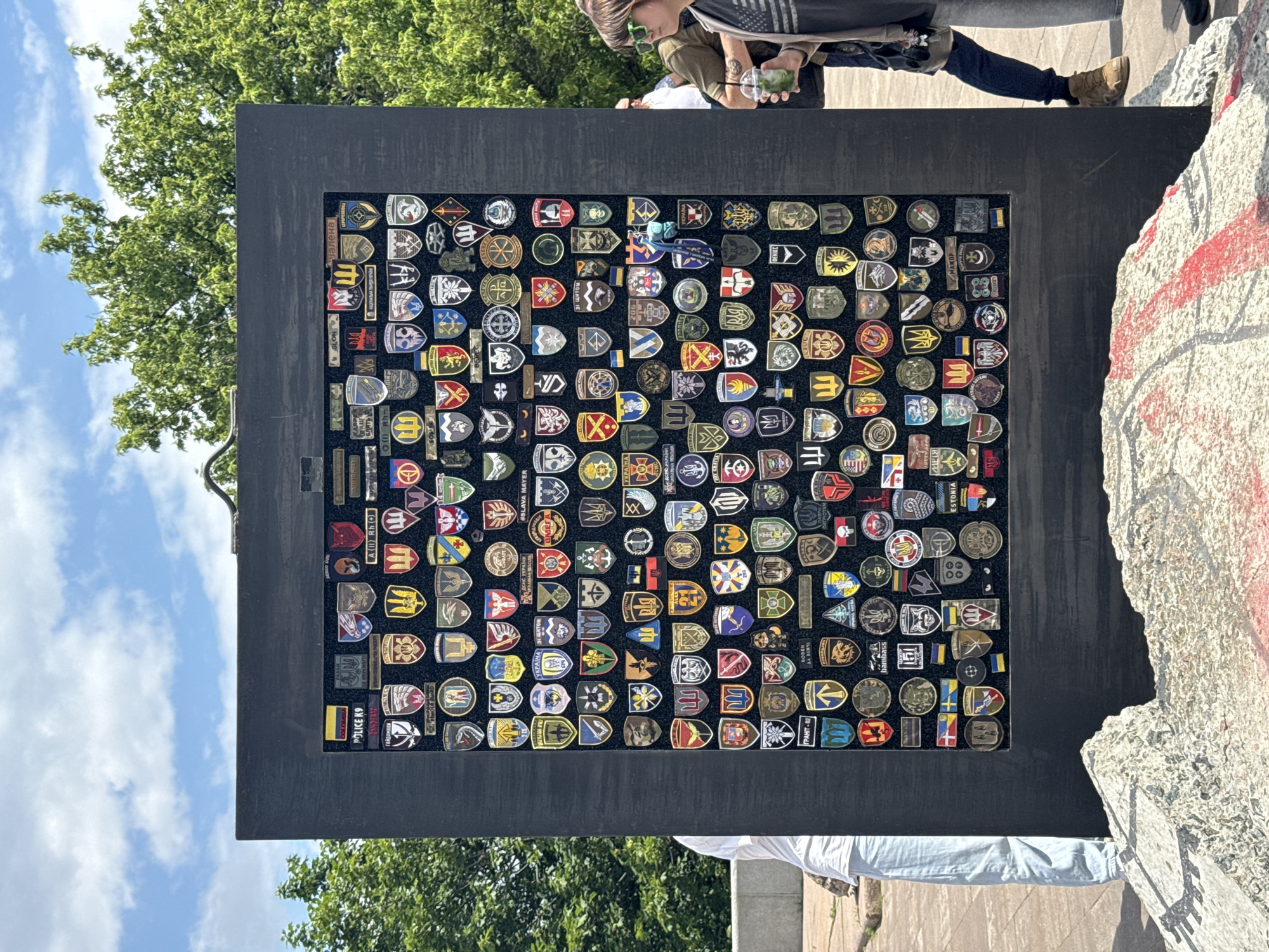 This is a medium-shot, vertical, eye-level photograph of a memorial display filled with various patches, likely military or law enforcement emblems, mounted on a black board. The display is located outdoors in Kyiv, Ukraine, as indicated by the context of the image and common knowledge of such memorials in the city. The memorial is positioned in what appears to be a public space, possibly a park or plaza, with lush green trees and a bright blue sky with scattered clouds visible in the background.
In the foreground, a portion of a large, rough, light-colored structure, possibly a concrete monument or a rock formation, is visible. This structure is marked with red paint streaks.
To the left of the memorial, a person wearing a white t-shirt and light blue pants is partially visible, standing with their back to the camera, looking at the display. To the right of the memorial, a woman wearing sunglasses and a dark jacket over a t-shirt, and dark pants, is holding a cup with a straw, also looking towards the display. Another person in a dark shirt and blue jeans is partially visible behind her, also looking in the same direction.
The memorial itself is rectangular and oriented vertically. It is densely packed with numerous patches of various shapes, sizes, and colors, each featuring intricate designs, symbols, and text, many in Ukrainian. The patches are arranged in a grid-like pattern. The black board on which they are mounted is framed by a thicker black border. The memorial appears to be a significant attraction or point of remembrance.