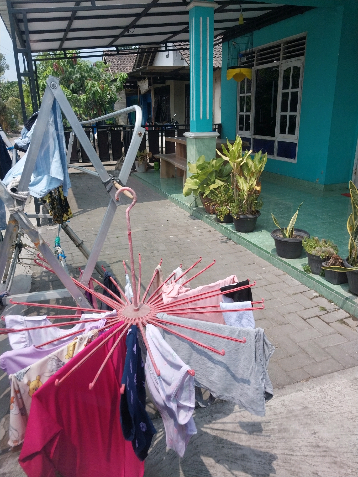 The image depicts a clothesline with laundry drying outside a turquoise-colored house. The clothesline is a pink, umbrella-like rack with various garments, including t-shirts, dresses, and pants, hanging from its spokes. Behind the clothesline, a silver metal clothes rack holds a light blue shirt and other items. The house has a tiled roof, white-framed windows, and a covered porch area with a wooden bench. Several potted plants are arranged along the front of the house. The scene appears to be set in an outdoor residential area with paved ground. The environment suggests a sunny day, as indicated by the shadows cast by the clothesline and rack. The specific location is Tirtomartani, Indonesia.