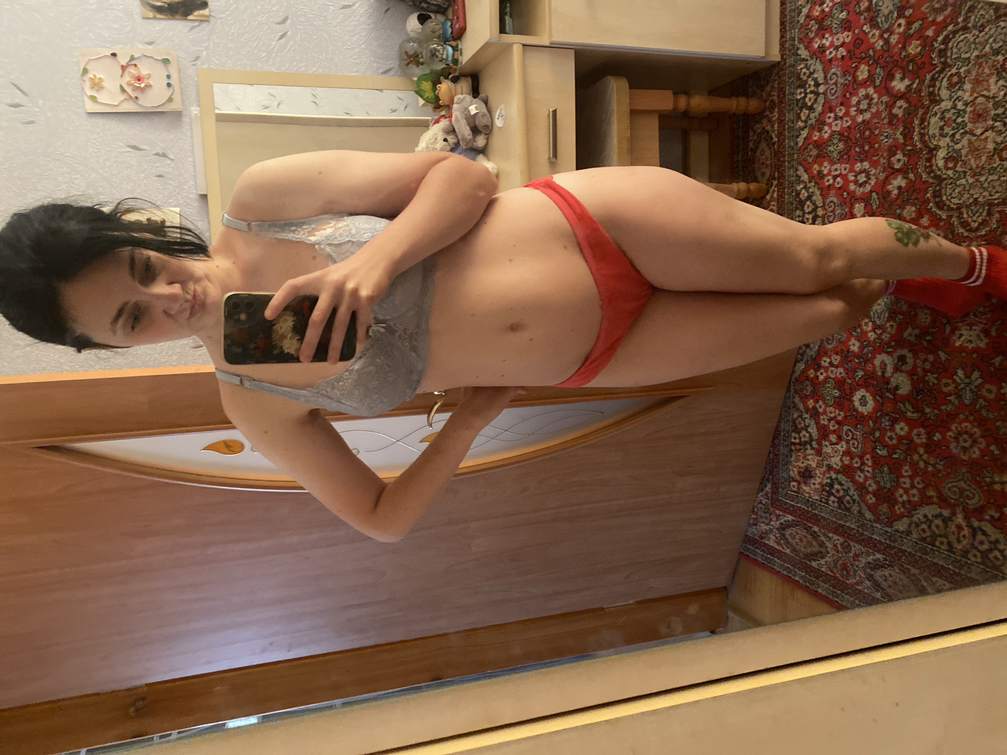 A woman is taking a mirror selfie in a room. She is wearing a grey lace bra and red underwear, along with red socks. She is standing in front of a wooden door and a vanity with a mirror. There is a rug on the floor.