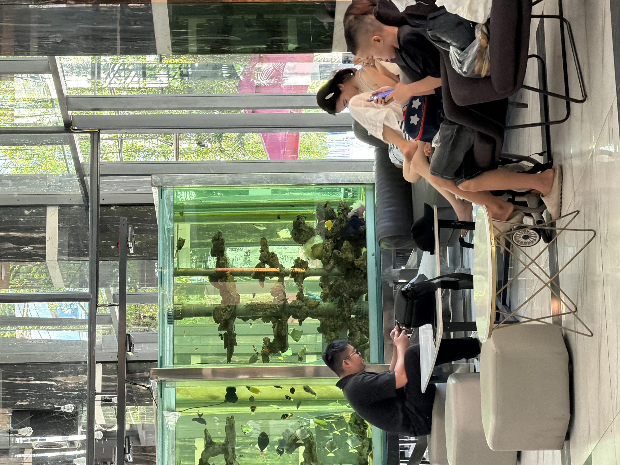 The image captures an indoor scene with a large aquarium filled with fish as the central focal point. The aquarium is situated behind a group of people who are seated and interacting with their mobile phones. In the foreground, a man in a black shirt is seated at a white table, looking down at his phone. To his right, a young woman with dark hair is also looking at her phone. To her right, a young boy in a black shirt is looking at his phone. The seating area is modern, with dark upholstered chairs and a dark sofa. In the background, large windows reveal an outdoor scene with lush greenery and trees, suggesting an urban environment. The architecture features elements of modern design, including large glass panels and metallic accents. The overall atmosphere is relaxed, with individuals engaged in their personal devices while in a shared public space. The presence of the aquarium indicates the location might be a hotel lobby, a restaurant, or a commercial establishment in Nha Trang, Vietnam, a coastal city known for its aquariums and modern architectural designs.