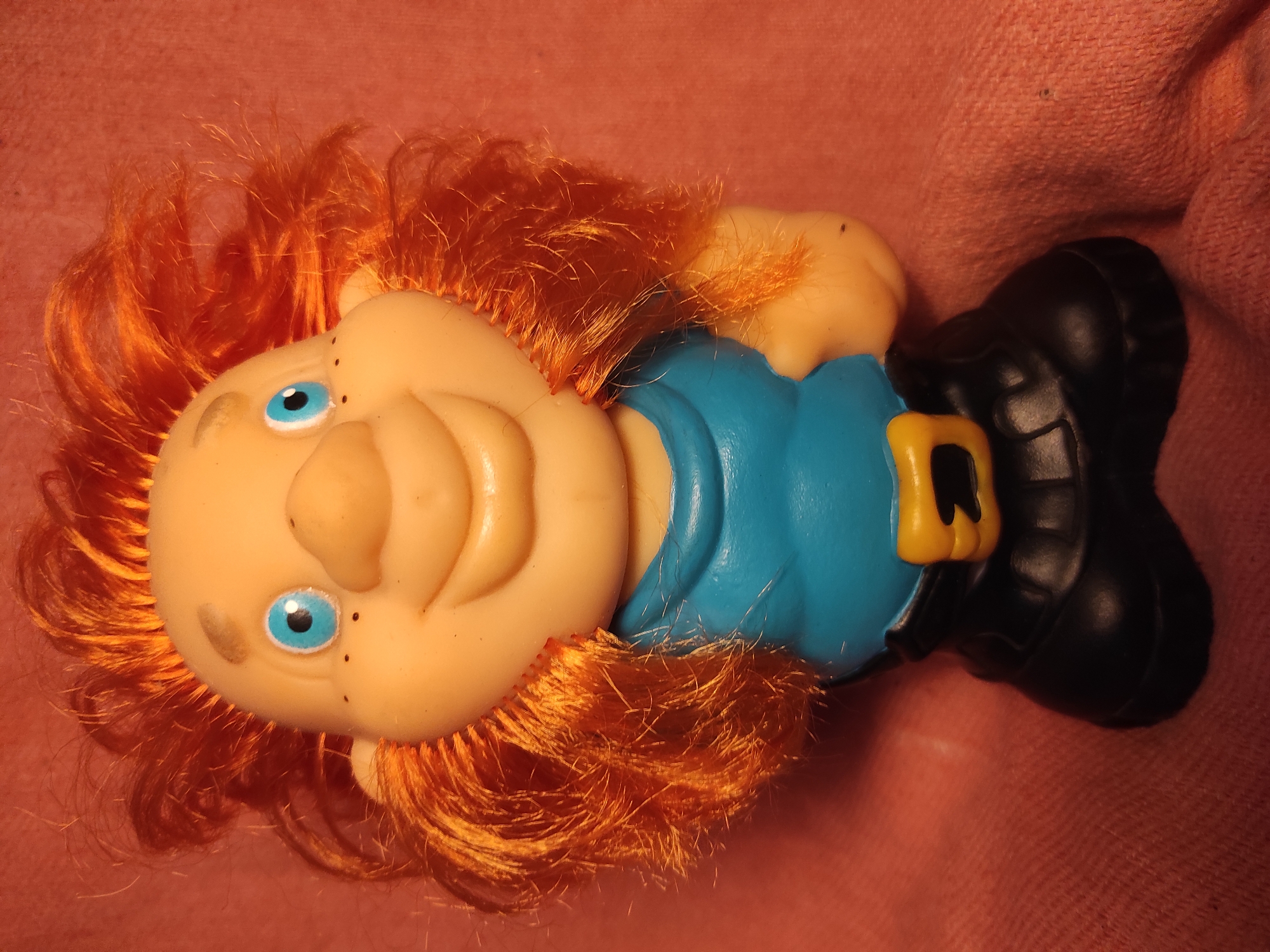 A plastic toy figure with orange hair, blue eyes, and a wide smile is pictured against a textured pinkish-orange background. The figure is wearing a blue shirt with a yellow belt and black pants. The figure's hair is styled in a wild, spiky manner, and it has small black dots on its cheeks, resembling freckles. The background appears to be fabric, possibly a blanket or tablecloth. The lighting is somewhat dim, casting shadows on the figure and background. No specific location in Chernihiv, Ukraine, can be identified from the image.