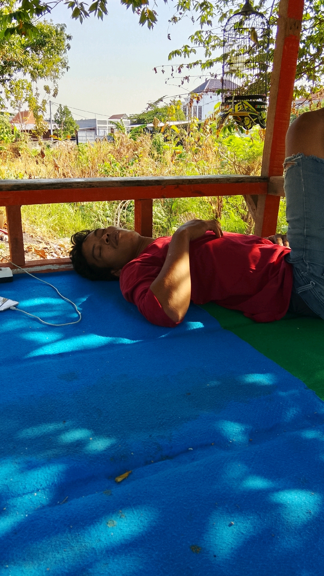 A man is sleeping on his back on a blue mat, under a wooden structure. He is wearing a red t-shirt and blue jeans. Sunlight filters through leaves, creating shadows on the mat. In the background, there is dry vegetation, buildings, and a birdcage hanging from the structure. The scene appears to be outdoors, possibly in a yard or park, in the city of Dahanrejo, Indonesia.