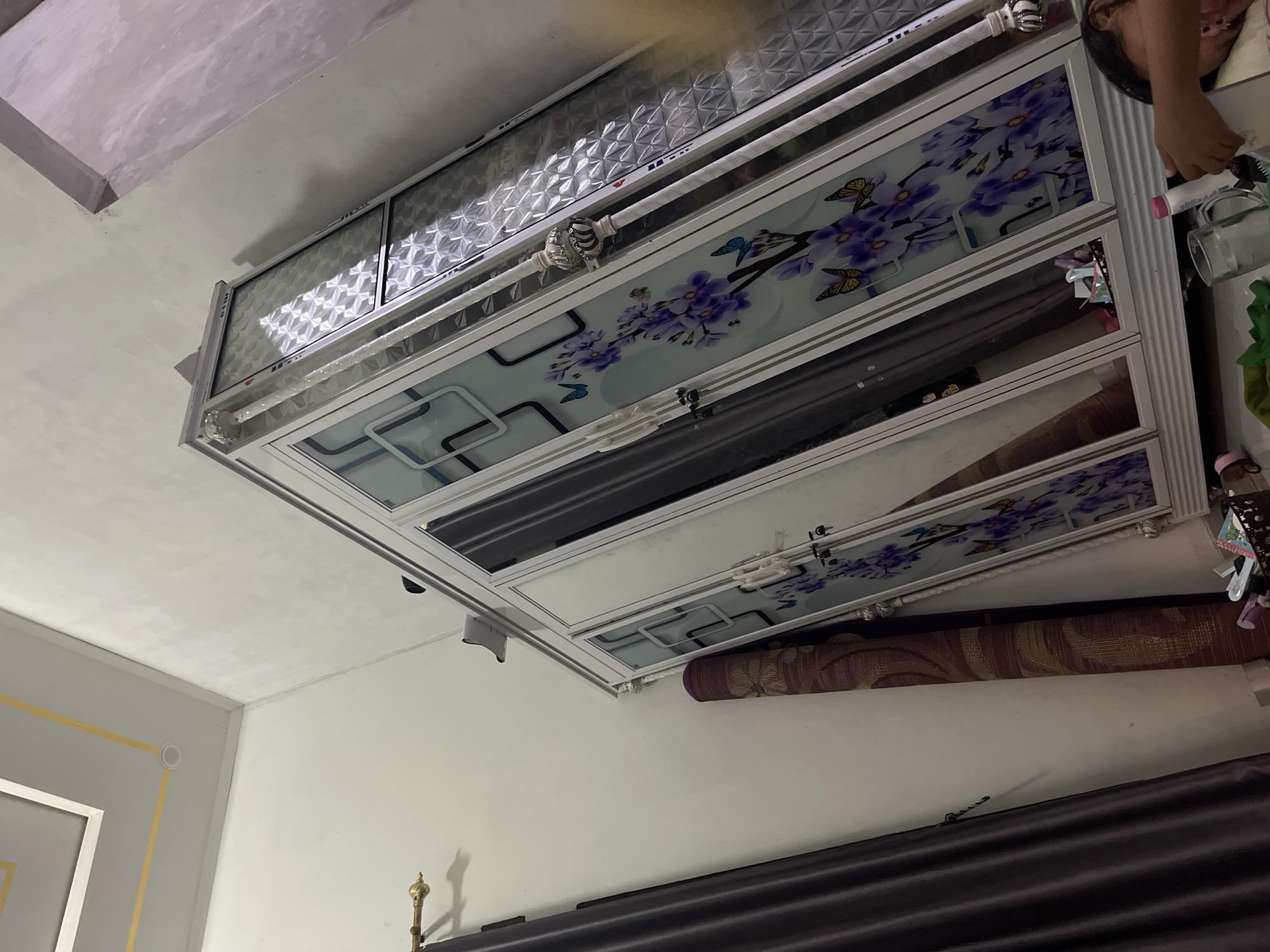 The image shows a bedroom in a residential setting in Kebumen, Indonesia. The primary subject is a large, white wardrobe with frosted glass doors decorated with floral and butterfly motifs. To the left of the wardrobe are dark purple curtains, and leaning against the wardrobe is a rolled-up rug with a brown and yellow pattern. The room has white walls and a white ceiling with yellow accents. In the lower right corner, a person's arm and hand are visible, holding a white bottle. A glass of water and other items are on the floor in front of the wardrobe. The room appears to be indoors, and the lighting suggests it is daytime.