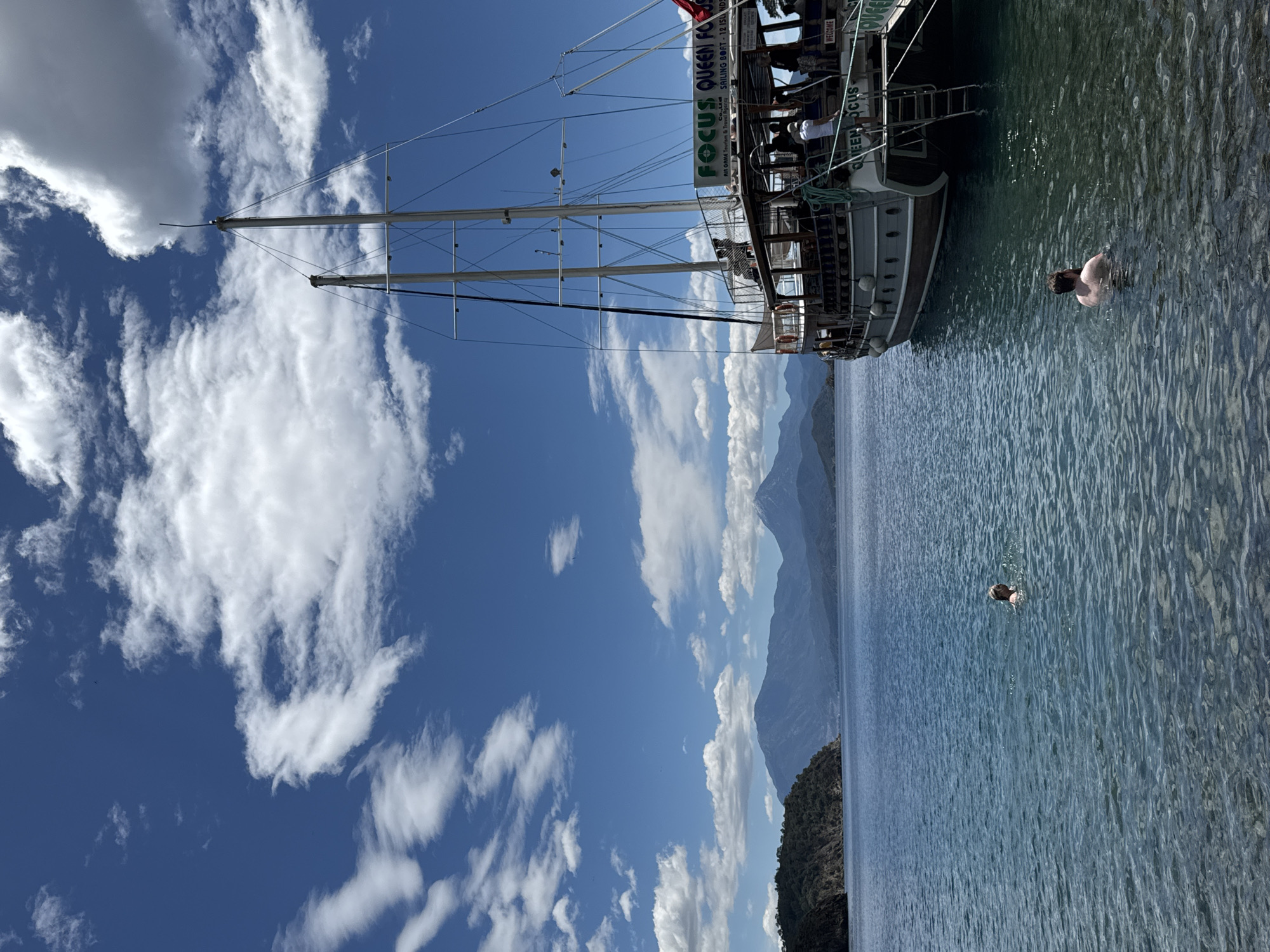 The image shows a boat, “FOCUS QUEEN FOCUS”, docked alongside a shoreline in Fethiye, Turkey. The boat is several stories high and is surrounded by water. A person is swimming in the water alongside the boat, and the masts reach toward the sky, partially obscuring the blue sky and white clouds. In the background, mountains are seen along the shoreline.