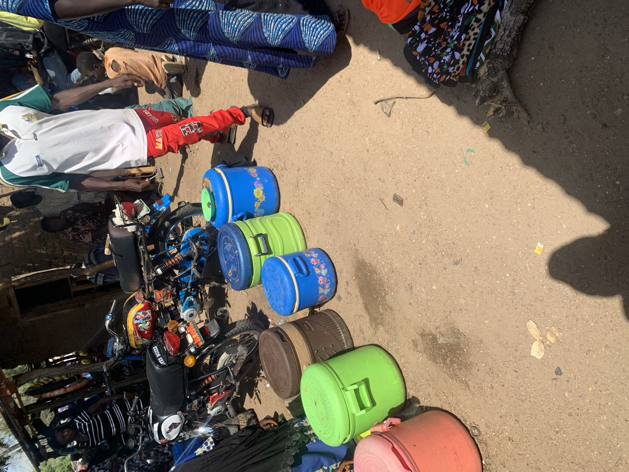 This image depicts a bustling scene outdoors, likely in Boi A, Nigeria, under the bright sun of a clear day. Several individuals are present, some of whom are engaged in activities related to a collection of objects. Prominently featured are several large plastic containers, in various colors including blue, green, and brown, some decorated with floral patterns. These containers appear to be either for sale or transport, as they are lined up on the ground.
Interspersed with the containers are motorcycles, suggesting a mode of transportation or perhaps a market setting where these items are sold. One person is seated on a motorcycle, dressed in a white shirt with green trim and red pants adorned with the number "57." Another individual, in an orange shirt, is leaning over, possibly interacting with the items or another person. Further back, a person in a black and white striped shirt is also near a motorcycle. The ground is a sandy, earthy surface, typical of an outdoor, possibly rural or semi-urban, environment. In the background, there are hints of structures and possibly more people and items, indicating a lively gathering.
The overall impression is one of commerce or daily life activity in a warm, sunny climate. The presence of people, motorcycles, and a variety of goods suggests a marketplace or a distribution point. The sunlight casts distinct shadows, confirming the time of day is likely midday. No overt emotions are discernible, but the arrangement of goods and people implies activity and interaction.