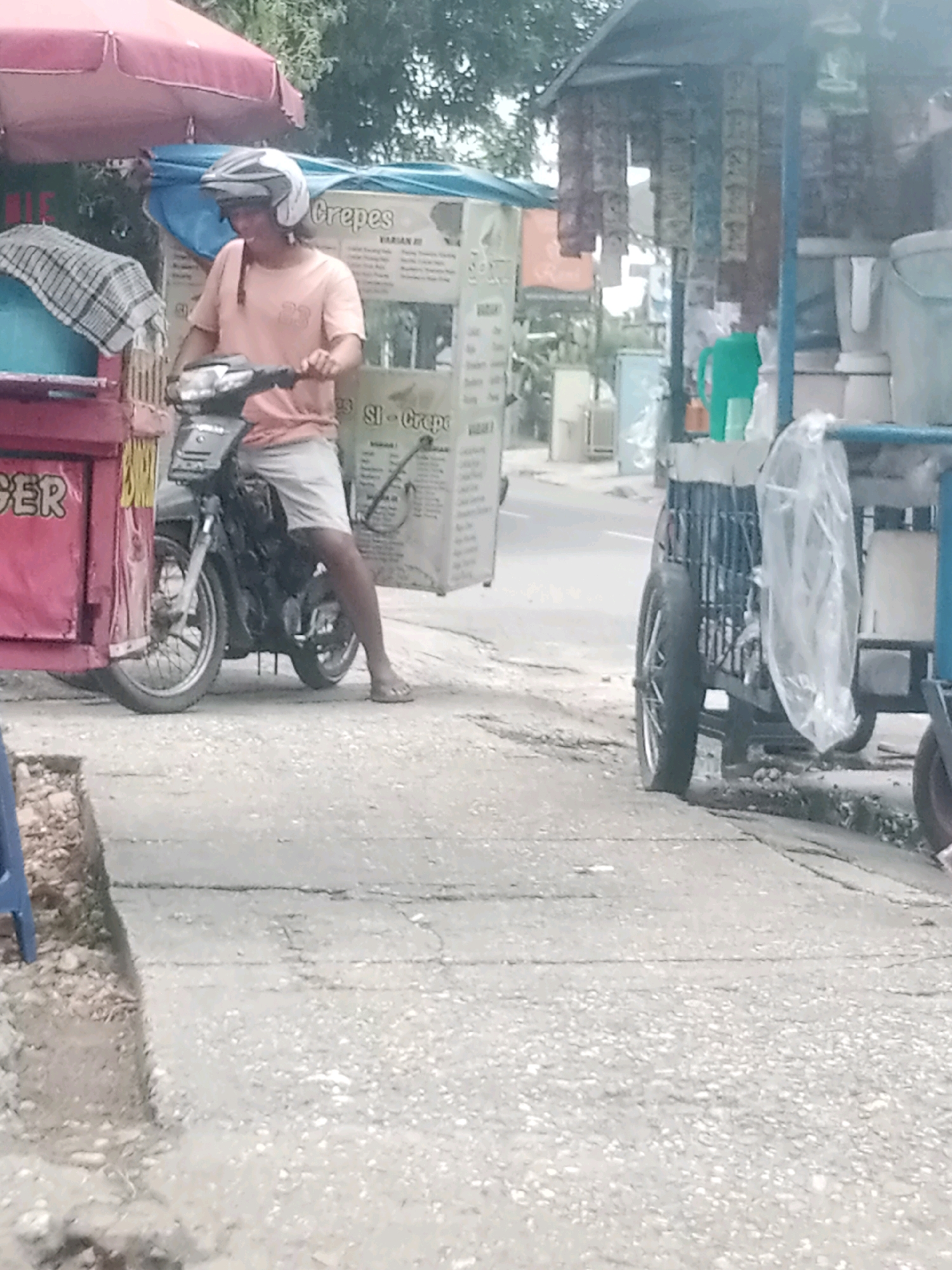 In Pekanbaru, Indonesia, a person wearing a helmet and an orange t-shirt is riding a motorcycle. The motorcycle is attached to a cart filled with boxes, likely for a food business, as indicated by the "Crepes" sign on one of the boxes. The person is operating the motorcycle near a roadside stall. The stall appears to be selling various items, including packaged goods hanging from the ceiling. The scene depicts a typical street vendor setup, with the cart and motorcycle acting as mobile vending units. The environment is outdoors, with a paved road and some vegetation in the background. The lighting suggests it is daytime.