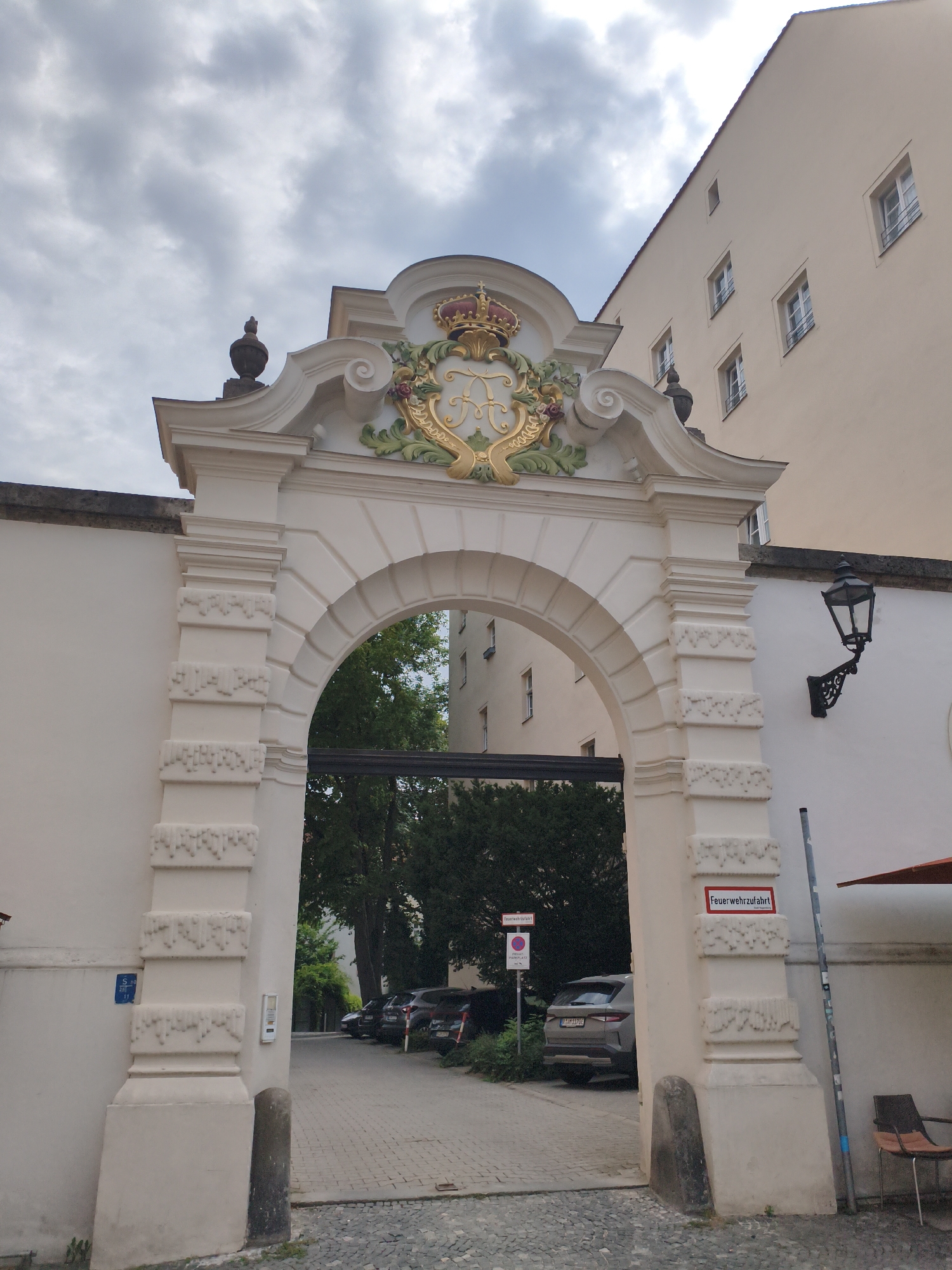 The image shows the historic Baroque gatehouse of St. Emmeram's Abbey in Regensburg, Germany. The gatehouse is made of white stone and features an ornate crest with a crown and the monogram "F" on top. Beyond the gate, a cobblestone driveway is visible, lined with trees and cars. The sky is cloudy. The scene is tranquil and appears to be a weekday. There are no people visible in the image. The architectural style of the gatehouse is elaborate and decorative, typical of Baroque architecture. The overall impression is one of historical significance and stately grandeur.
