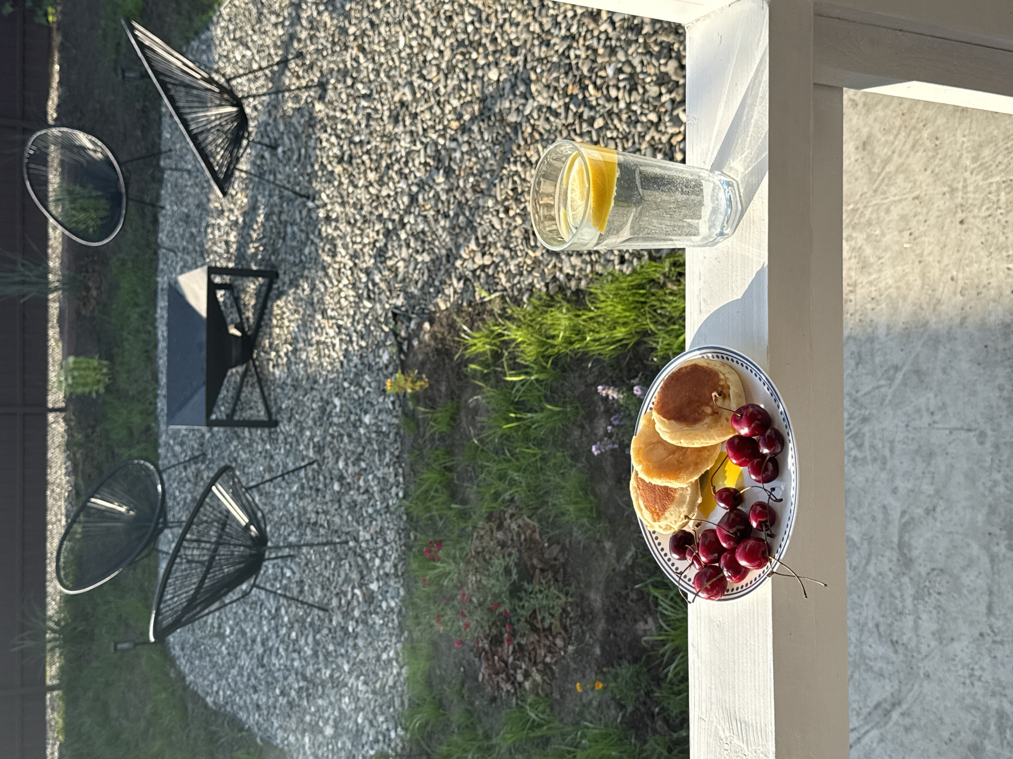 In the city of Starokorsunskaya, Russia, an outdoor scene is depicted. In the foreground, a white railing supports a plate of three golden-brown pancakes and a pile of dark red cherries, alongside a tall glass of clear liquid with lemon slices. The plate and glass are positioned on the right side of the frame. Behind the railing, a gravel patio is visible, featuring a dark square fire pit in the center, surrounded by four black Acapulco chairs with woven seats. Beyond the patio, a patch of green grass and a small garden area with red and yellow flowers are present. The scene is bathed in the warm light of the setting sun, casting long shadows. The background shows a dark wooden fence and more green foliage.