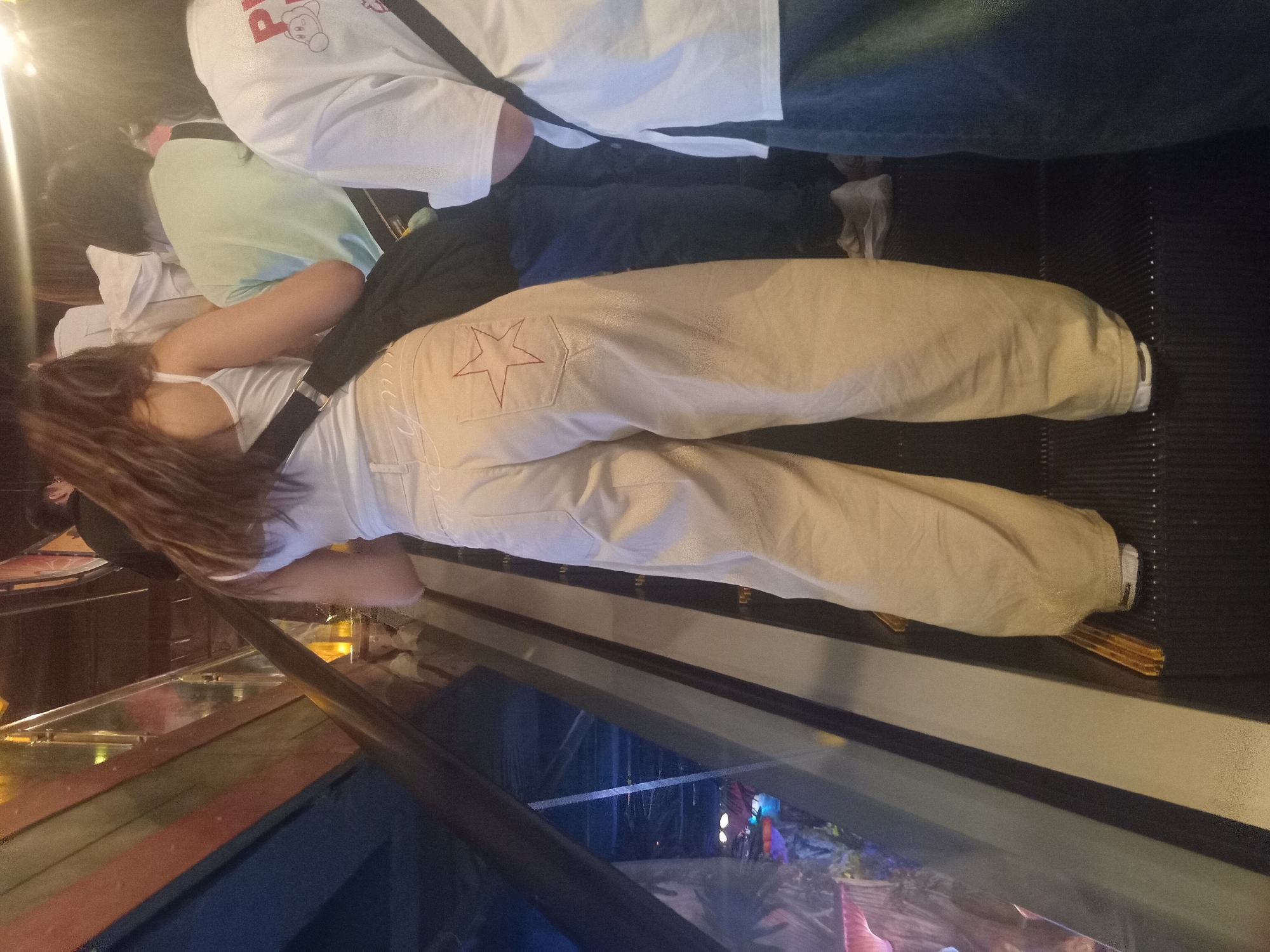 The image shows a woman from behind on an escalator, wearing a white tank top and beige pants with a red star design on the back pocket. She is carrying a black bag across her chest. Behind her, other people are also on the escalator. The environment appears to be an indoor public space with wooden and metallic structures. The scene takes place on an escalator in the city of Đà Nẵng, Vietnam.