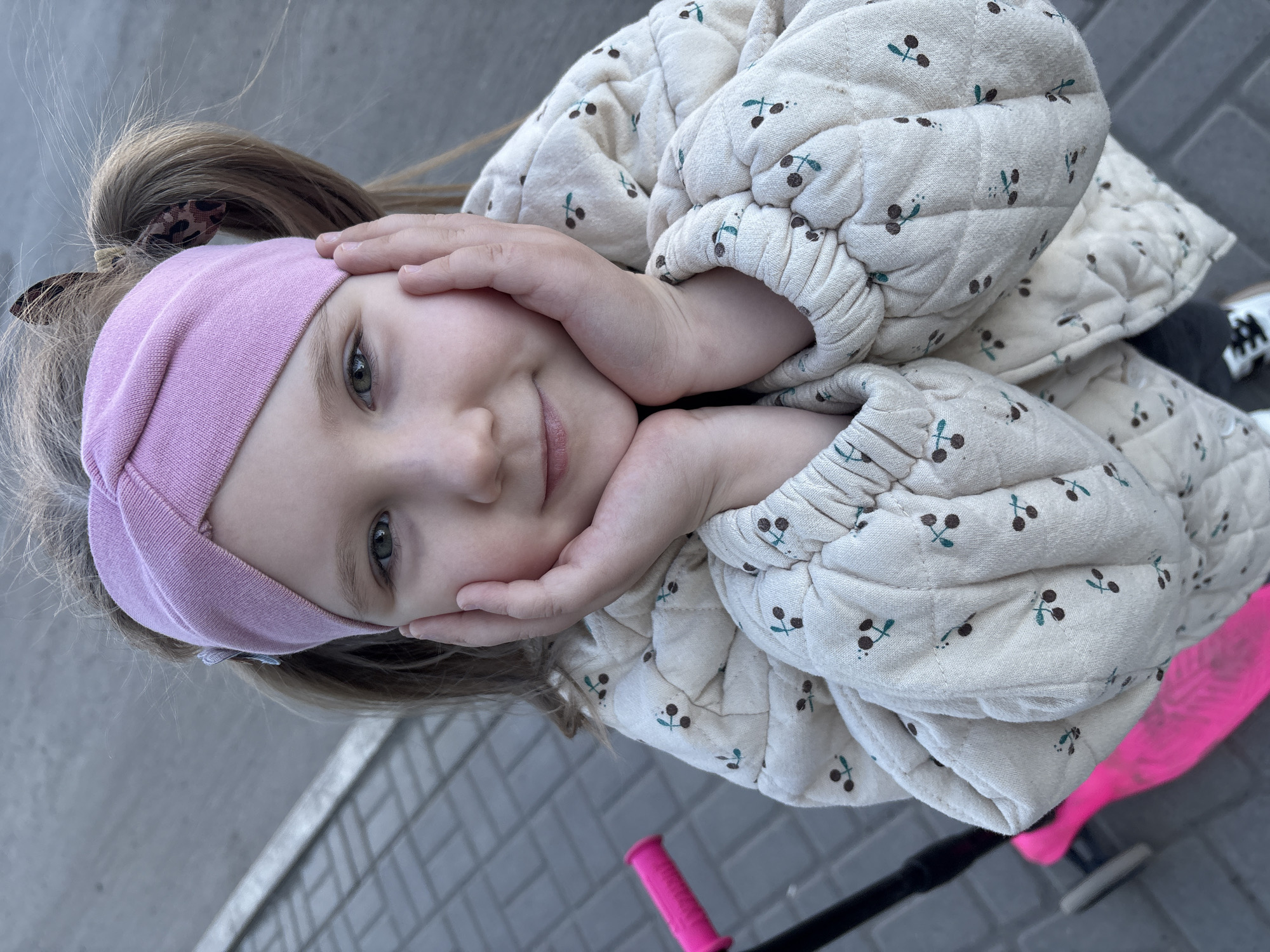 This brightly lit, slightly tilted, portrait-style photograph captures a young child outdoors in Chaikovsky, Russia. The child, likely a girl with light brown hair pulled back into a ponytail, looks directly at the viewer with a gentle, slight smile. She wears a soft pink headband across her forehead and a cream-colored quilted jacket patterned with small dark cherries and green stems. Her hands are positioned on either side of her face, with fingers lightly pressing against her cheeks, enhancing her thoughtful expression. Her eyes are a striking grey-green.
The background features a blurred, neutral grey tone, possibly a wall or building facade. The ground below is paved with grey rectangular stones, suggesting an urban or suburban outdoor environment. Partially visible in the lower right corner is the bright pink handle and a portion of a scooter, indicating a recreational activity. The overall impression is one of calm and innocence, captured in natural daylight. There is no visible text in the image.