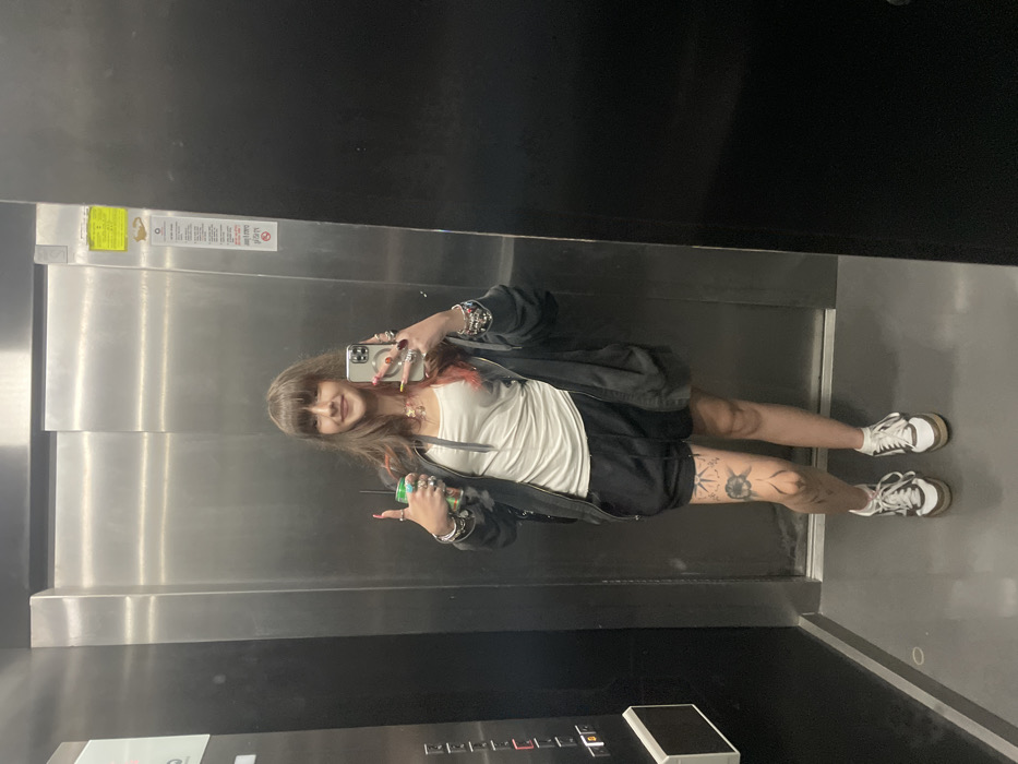 Inside a mirrored elevator, a woman takes a selfie. She wears a white tank top, black shorts, and a dark jacket. Her left leg features a tattoo. She holds a green can in her left hand and her phone in her right. The elevator is metallic, reflecting the scene. A yellow sticker is visible near the top of the elevator wall. Buttons are at the bottom.