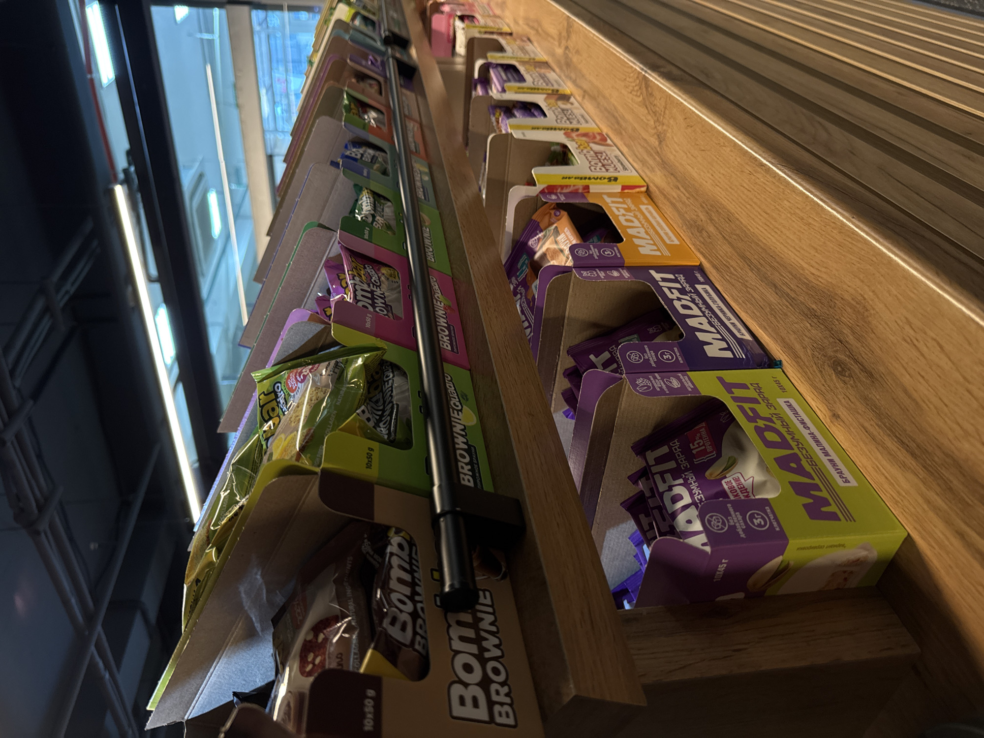 Here's a forensic description of the image:
**Scene:**
The image shows a display shelf stocked with various packaged brownie snacks. The shelf is made of wood, with a light brown and dark brown grain pattern. A black metal bar runs horizontally along the front edge of the shelf, likely to prevent items from falling.
**Objects:**
* Packages of "Bombbar Brownie" snacks are prominently featured, with the "Bombbar" logo visible.
* Packages of "Madfit" brownies can also be seen in yellow, orange, and purple packaging.
* The packages are oriented to face the viewer, arranged in a row on the shelf.
**Environment:**
The scene appears to be inside a store or shop. Bright light is visible through a window or opening in the background.
**Location (implied):**
The packaging includes the words "БРАУНИ МАЛИНА-ФИСТАШКА 10Х45 Г" in Russian, which translates to "Brownie Raspberry-Pistachio 10x45 g". The packaging also includes Russian text in other sections. This strongly indicates that the store is located in a place where the Russian language is prevalent. Sochi, Russia is specified, therefore, it is most likely in a store in Sochi, Russia.