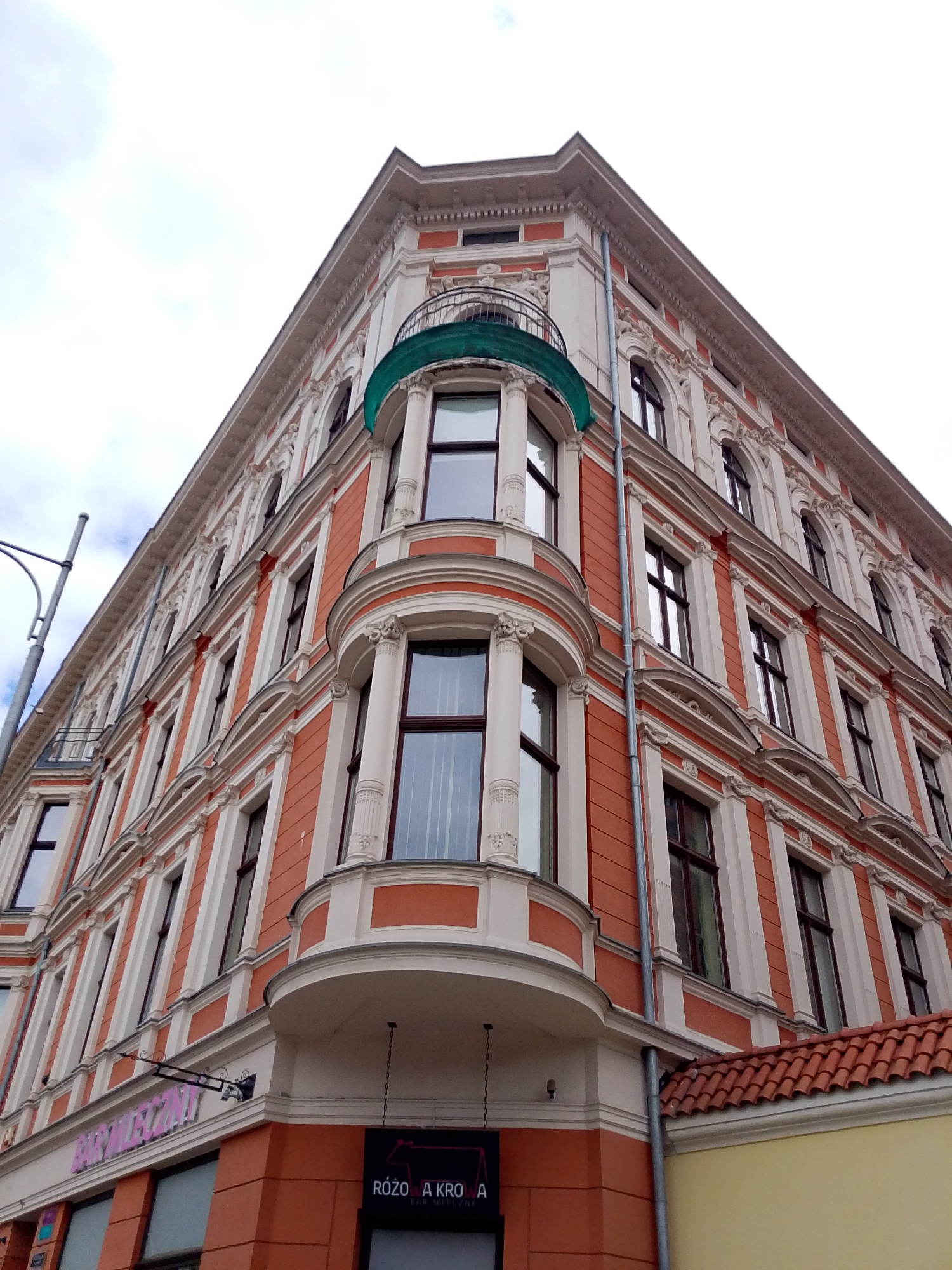 This is a low-angle, eye-level photograph of the corner of an ornate, historic building in Wrocław, Poland. The building has a salmon-orange facade with white decorative elements around the windows and along the horizontal cornices. The building has multiple stories with arched windows on the upper floors and rectangular windows on the lower floors. A curved balcony with a green canopy juts out from the corner of the building on the second floor. Below the balcony, there is a sign with pink lettering that reads "BAR MLECZNY" and another sign with a black background and white text that appears to read "RÓŻOWA KROWĄ". The building is situated on a street, as indicated by a street lamp on the left side of the frame and part of a tiled roofline on the right. The sky above is overcast and pale.