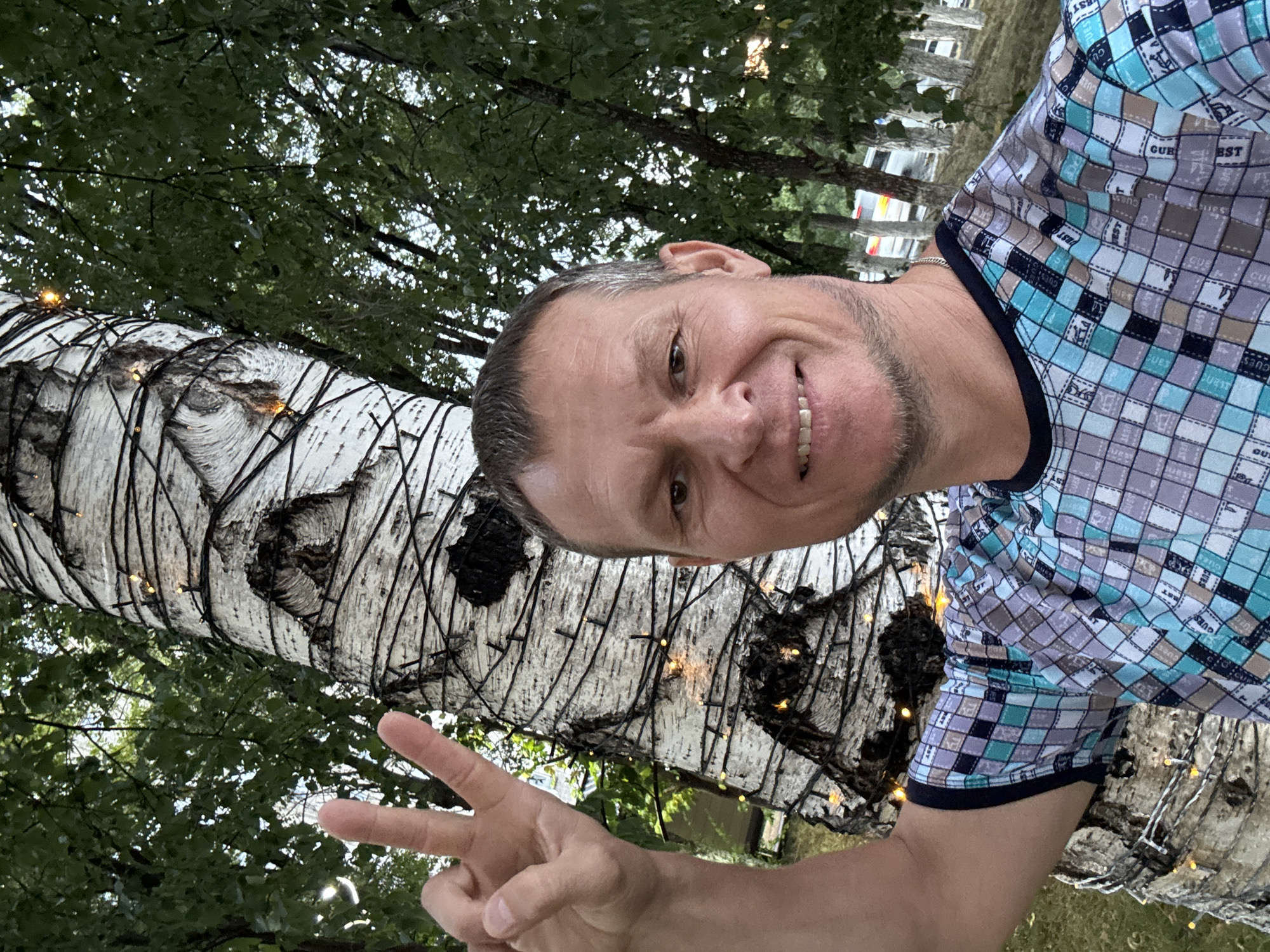 A man in a checkered t-shirt smiles at the camera, holding up two fingers in a peace sign. He stands in front of a birch tree wrapped with string lights. In the background, there are green trees, and a few parked cars are visible beyond them. The location is Oskemen, Kazakhstan.