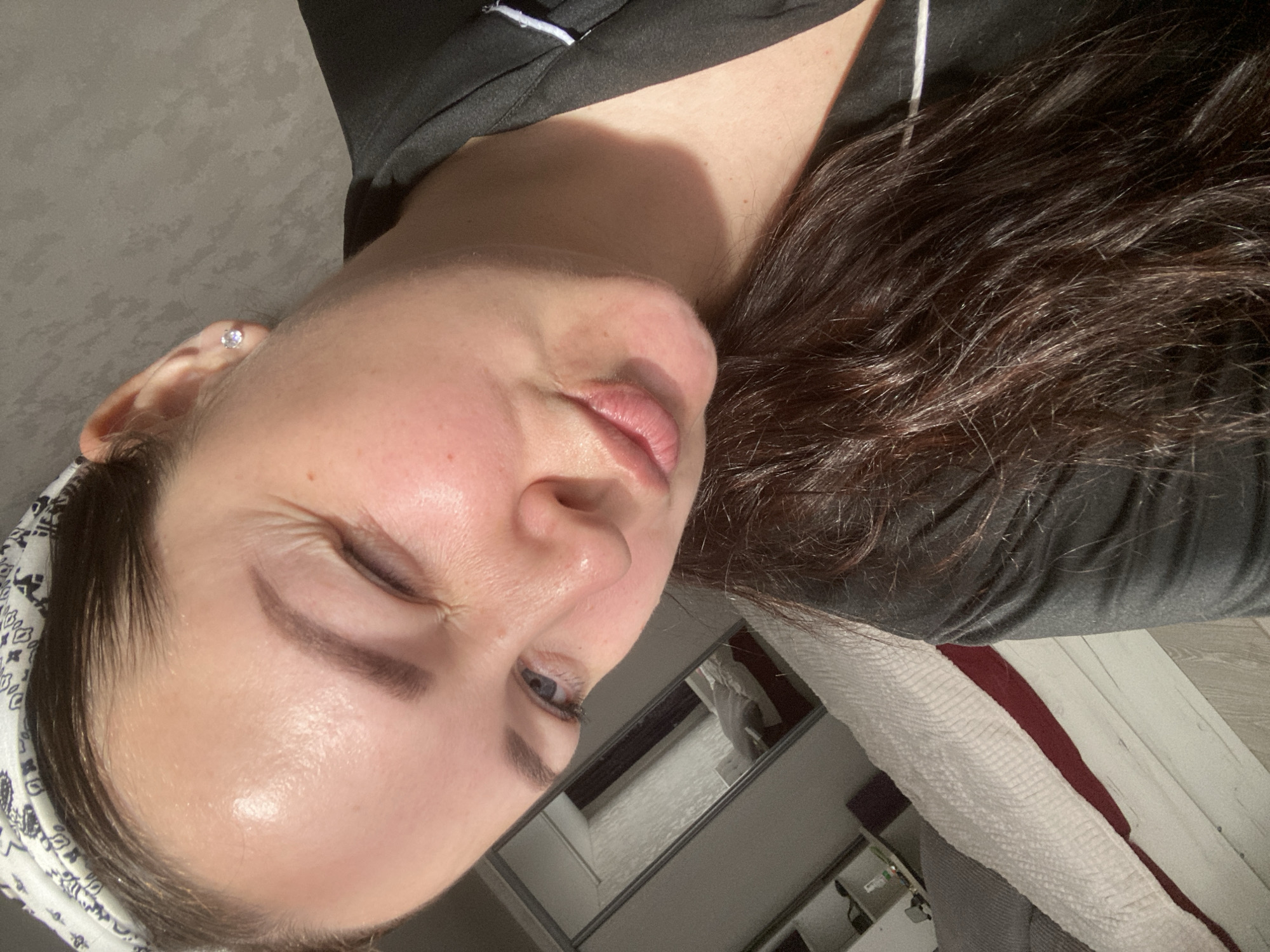 A close-up, rotated image shows a woman's face. She is fair-skinned with dark brown hair. She is wearing a dark-colored shirt with a light trim and a patterned head covering. A small earring is visible in her left ear. Her eyes are partially closed. The background includes a mirror reflecting a room with white and maroon bedding. The location of the image is not discernable from the given information.