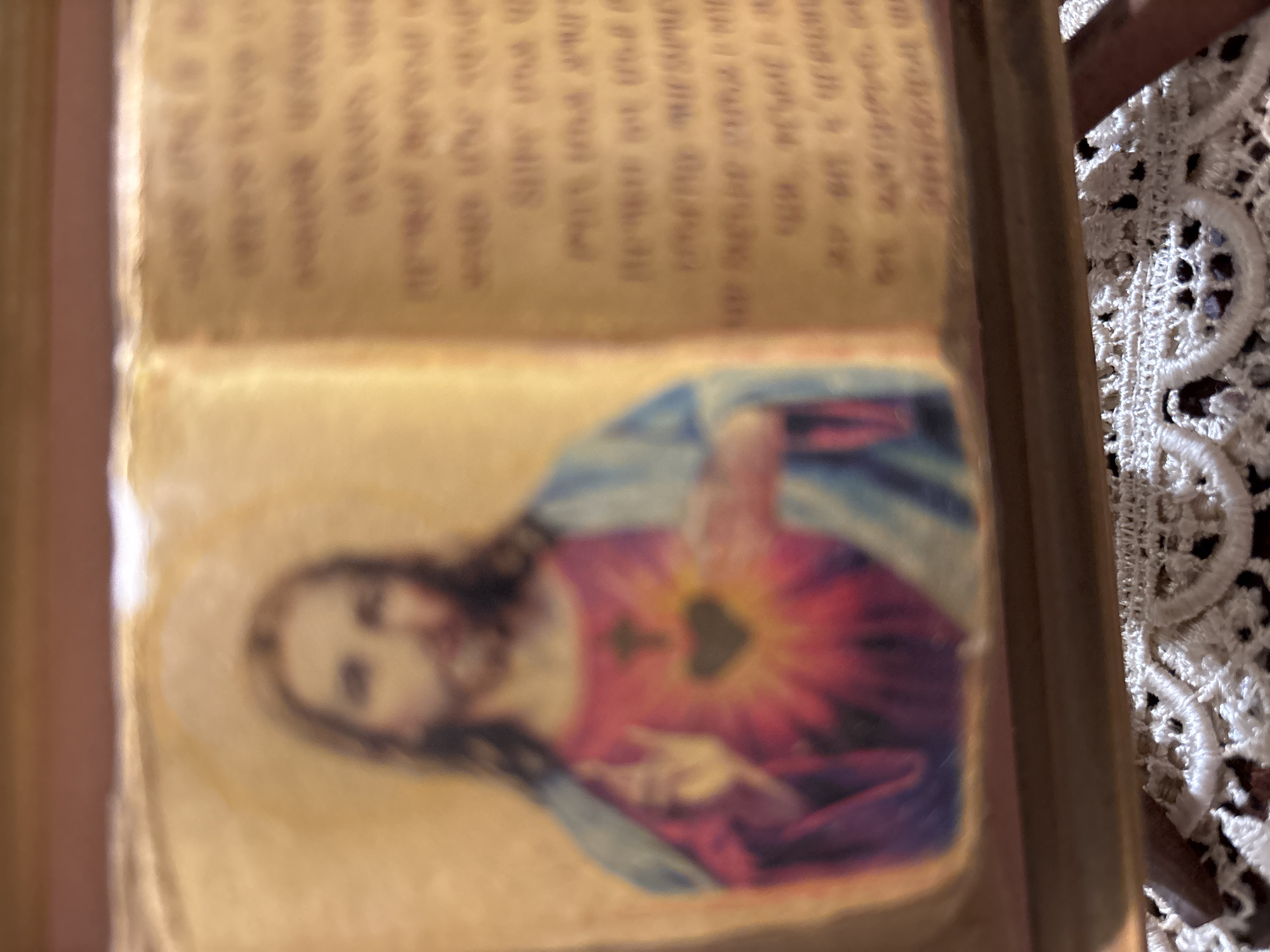 The image presents a blurry, close-up view of an open book, displaying a religious illustration on one of its pages. The central subject is a depiction of Jesus Christ, rendered in a colorful style, affixed or printed onto the right-hand page. Jesus is shown with long dark hair, a beard, and a subtle halo, wearing robes that appear to be blue and red/purple. His right hand is raised, and his chest prominently features the Sacred Heart, an iconic symbol of Jesus's divine love, depicted as a flaming heart radiating light.
Above this illustration, on both the right and left pages of the open book, there is printed text. However, the entire image is significantly out of focus, rendering all visible text completely illegible. The book pages have a warm, aged, yellowish-brown tone, suggesting an older volume.
To the right edge of the book, a section of white, intricately patterned lace fabric is visible, possibly part of a tablecloth, altar covering, or other decorative textile. The background is indistinct and softly blurred, contributing to a serene atmosphere. The scene evokes a sense of devotion or contemplation, likely taking place indoors, with the provided context indicating a location in Vagharshapat, Armenia, though no specific visual cues within the image confirm this.