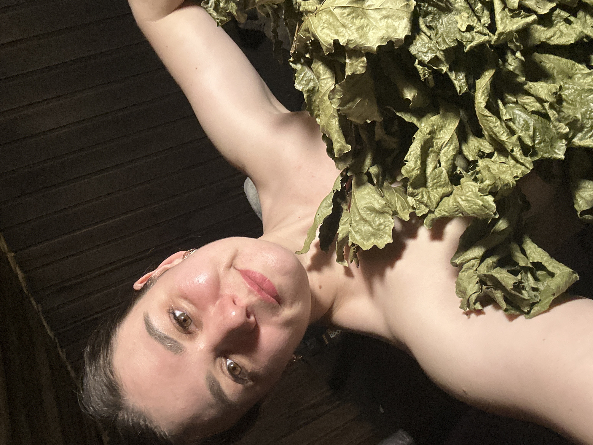 A person, presumably a female, is holding a bunch of dried leaves. The background appears to be wooden planks, possibly the interior of a sauna or bathhouse. The location is speculated to be in the city of Chaikovsky, Russia.