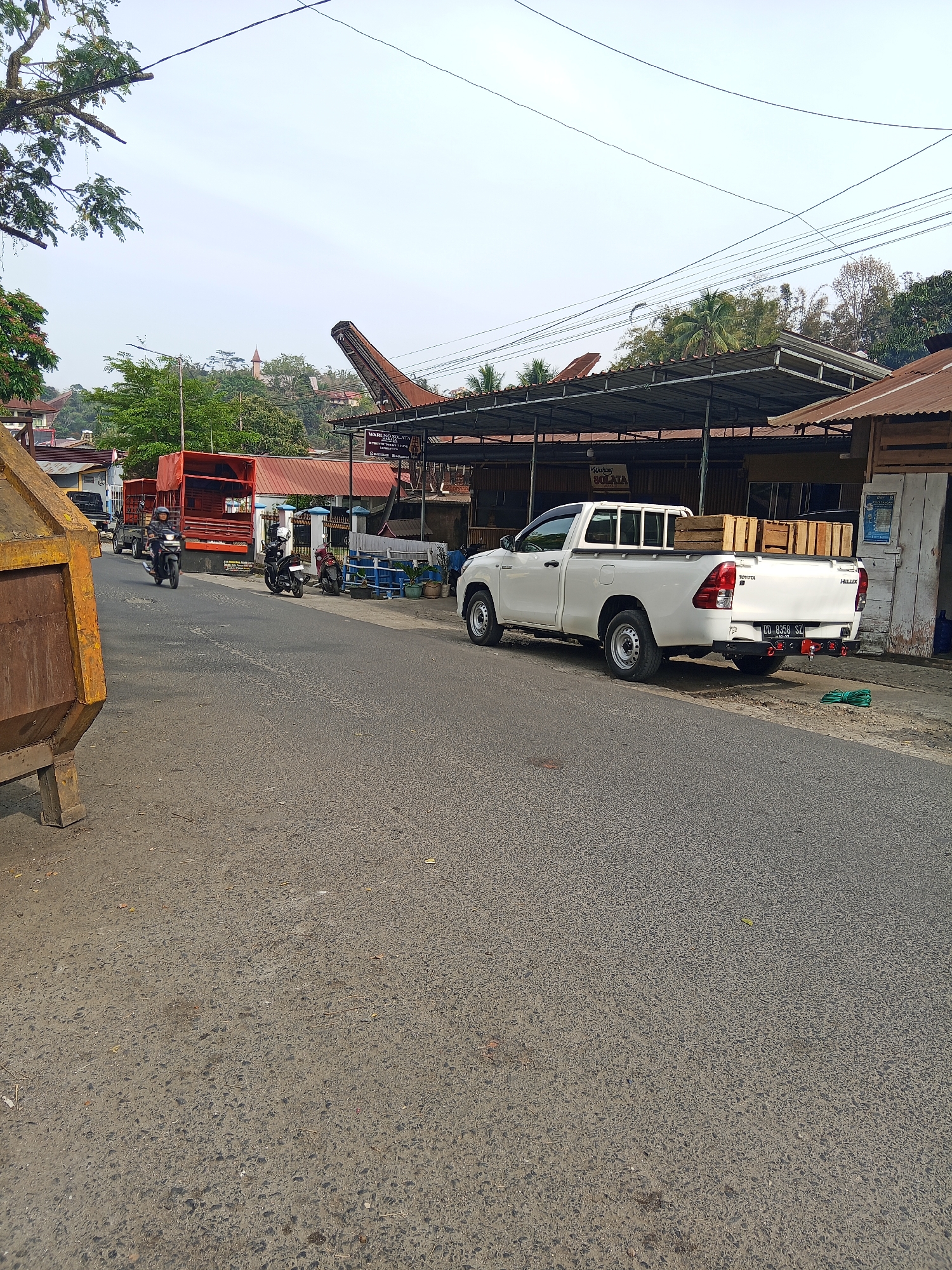 A street scene in Makale, Indonesia, shows a white Toyota Hilux pickup truck loaded with wooden crates parked on the right side of the road. The truck is facing away from the camera. In the background, a covered outdoor seating area with tables and chairs is visible. A red flatbed truck is driving away from the camera on the left side of the road, with two motorcycles following behind it. A large, rusty dumpster is in the foreground on the far left. The sky is overcast, and there are power lines crisscrossing above. The environment appears to be a commercial or roadside area with some residential buildings and vegetation on a hillside in the distance.