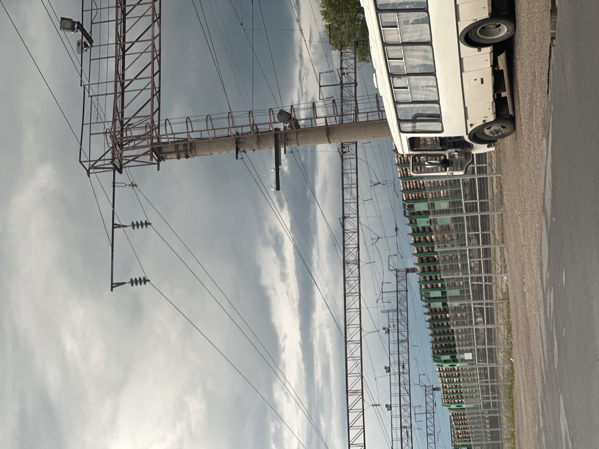 An outdoor, eye-level, medium shot captures a cloudy day in Plesetsk, Russia, at what appears to be a train station or industrial area. In the foreground, a section of asphalt with a puddle is visible. To the right, the rear half of a white bus is parked, its door open and curtains drawn inside. Behind the bus, a green fence separates the immediate area from a train track. The train track is lined with a train car filled with logs. Overhead, a complex network of power lines and metal structures, including a ladder attached to a concrete pole, crisscross the sky. The sky itself is heavily overcast with dark clouds.