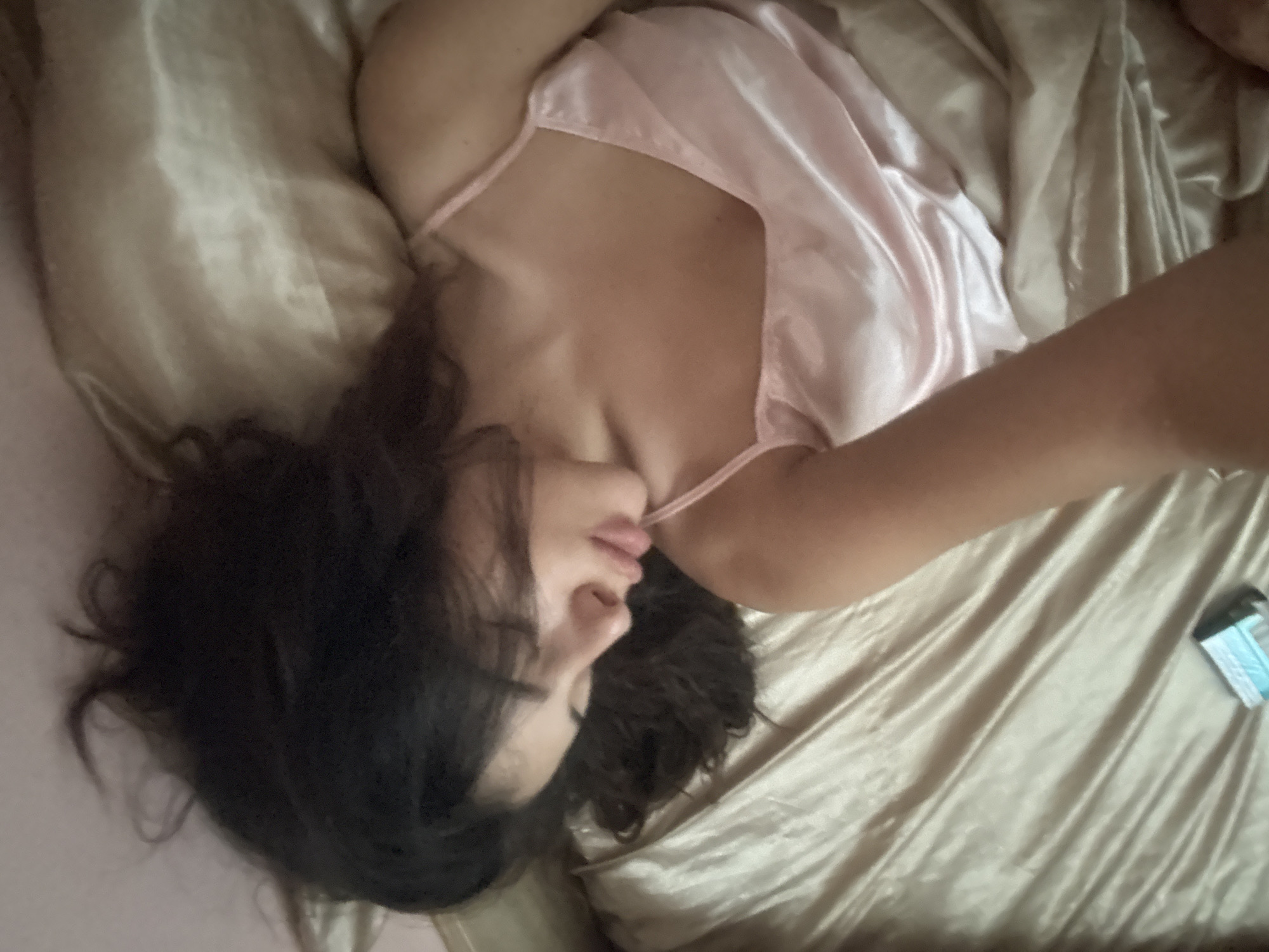 The image shows a woman lying in bed, covered by satin sheets. Her eyes are closed, and she appears to be sleeping. A small, rectangular object, possibly a pack of tissues or a medication box, is visible on the left side of the bed. The sheets are a light, shimmery gold color, and the woman is wearing a pink slip. The scene is lit by soft, natural light. The image does not contain any identifiable landmarks or elements that would indicate a specific location in Plovdiv, Bulgaria.