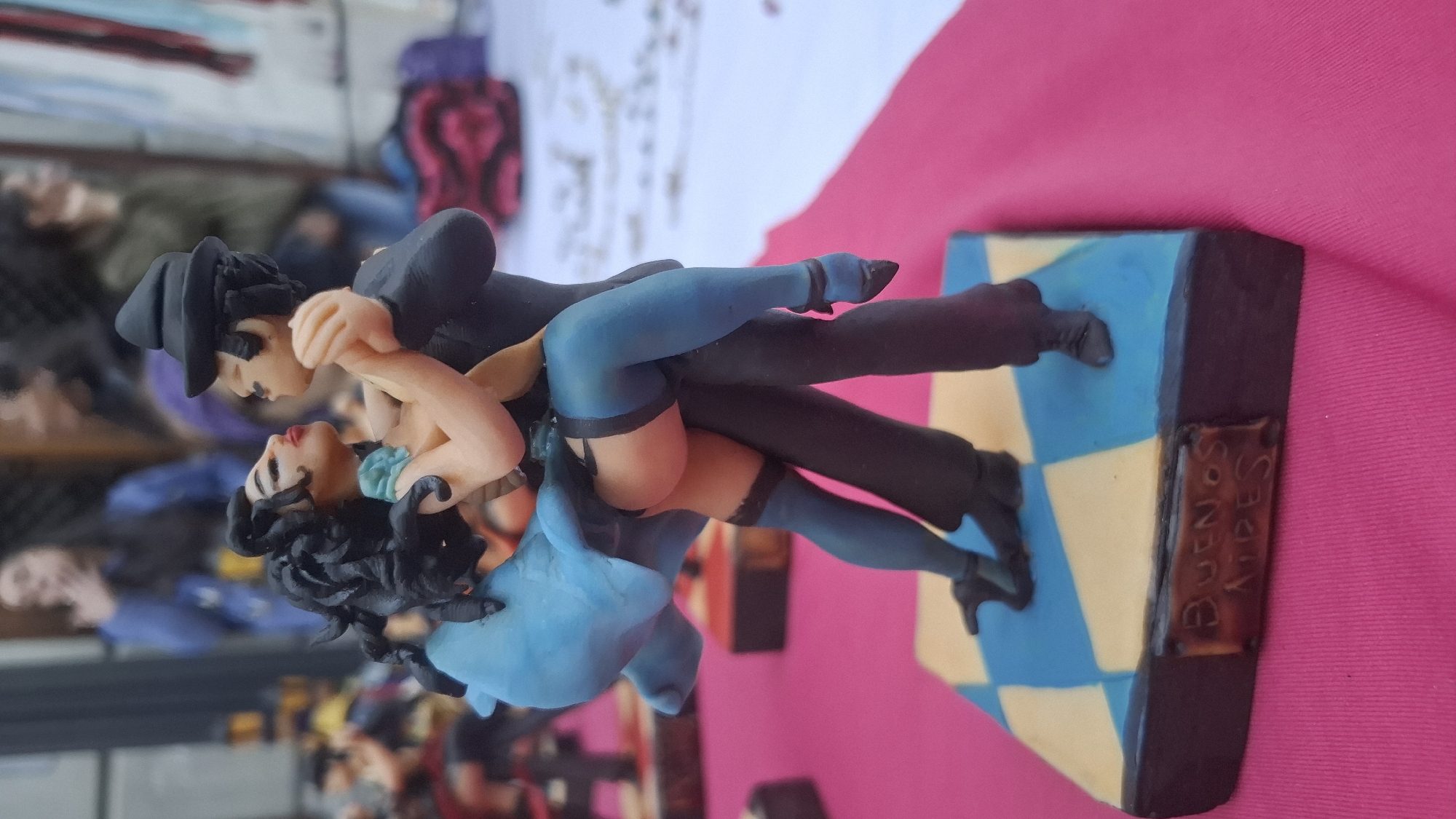 A figurine depicting a couple dancing the tango stands on a blue and yellow checkerboard base labeled "Buenos Aires". The man wears a black suit and hat. The woman has black hair and wears a blue dress and blue stockings. The background is blurry but suggests an outdoor setting with people and objects.
