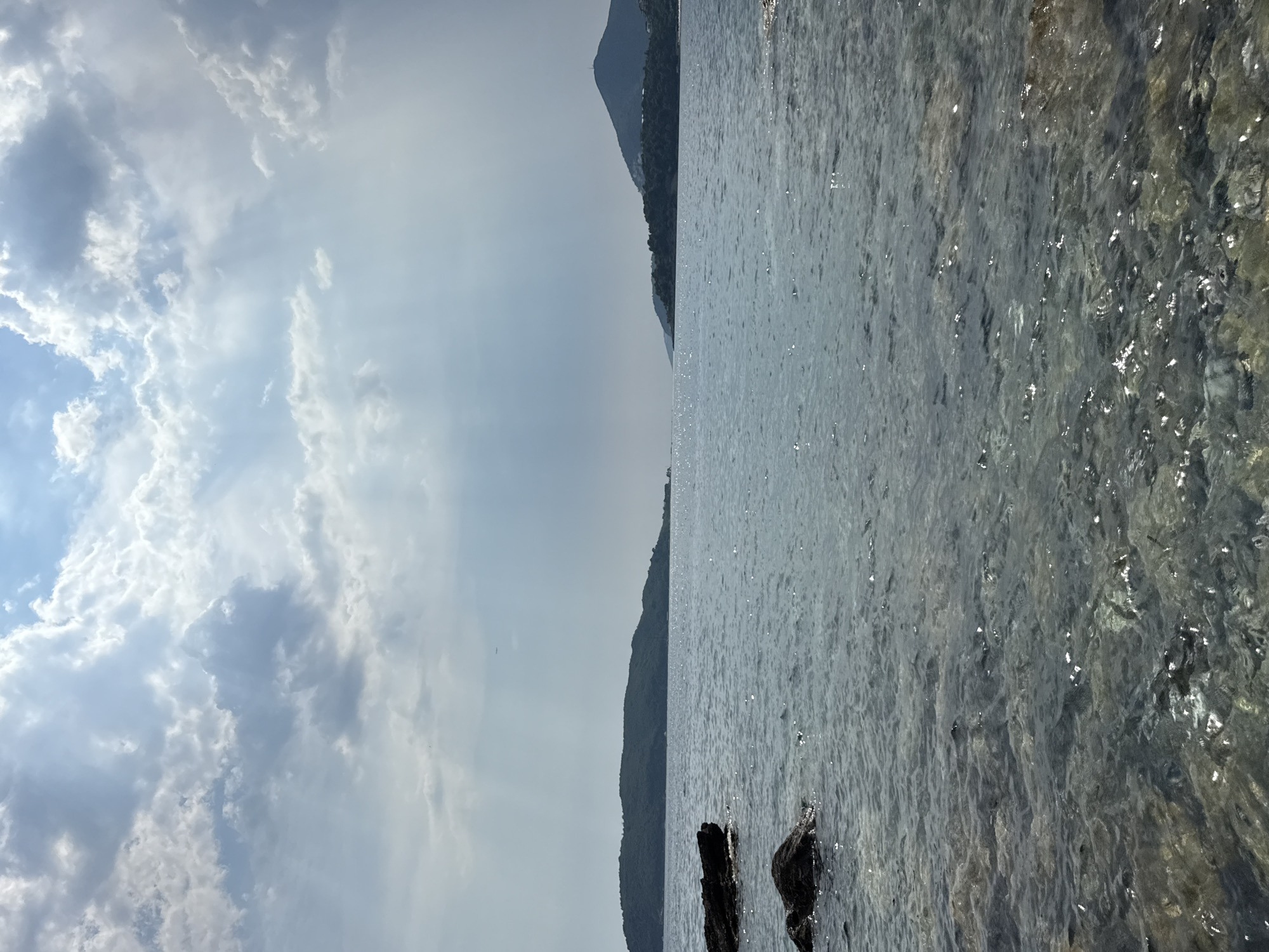 This media file displays a scenic view of a body of water with a mountainous landscape in the background, likely Bečići, Montenegro. The image is oriented vertically, with the sky occupying the left half and the water and land on the right.
The sky is filled with a dramatic display of clouds, ranging from bright white to darker grays, suggesting dynamic weather conditions. Sun rays are visible, piercing through some of the clouds, indicating it might be daytime, possibly late afternoon or early morning when the sun is lower in the sky.
The water appears to be a calm sea or lake, with light reflecting off its surface, creating a shimmering effect. The clarity of the water is noticeable, with some submerged rocks or the seabed visible in certain areas. Two dark, weathered pieces of driftwood or rocks are partially submerged in the foreground of the water.
The background features a sloping, verdant mountainside, heavily forested, with a peak visible in the distance. The silhouette of the mountain range against the hazy sky suggests a sense of depth and scale. The overall mood of the image is peaceful and natural, evoking a sense of tranquility and the beauty of the coastal environment. No people or text are visible in the image.