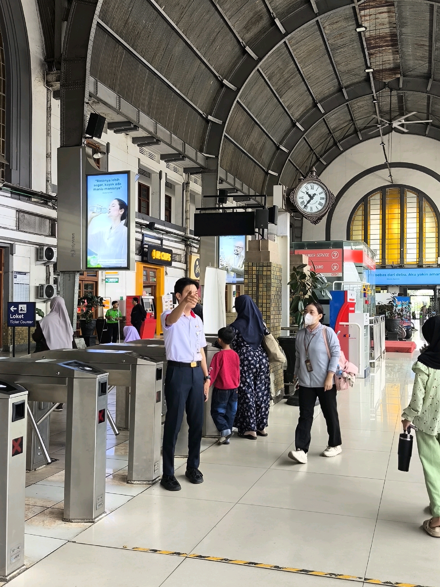 The image depicts the interior of a train station in West Jakarta, Indonesia, specifically at Gambir Station, on a bright day. The station features a high, arched, corrugated metal roof supported by dark beams. Sunlight streams through large, arched windows with yellow-tinted glass at the far end of the hall. A large, ornate clock with Roman numerals is mounted on the wall overhead.
In the foreground, a man in a white short-sleeved shirt with a red collar and a dark tie, dark pants, and black shoes stands near ticket gates. He is gesturing with his right hand, appearing to give directions. Several ticket gates, made of brushed metal with red indicator lights, are visible.
Several individuals are present in the midground and background. To the left of the ticket attendant, a person wearing a light grey headscarf and a black top is partially visible near a ticket counter sign. Further left, a large digital display shows an advertisement with an image of a woman drinking water and text in Indonesian.
Other people are moving through the station. A woman wearing a dark blue hijab and a patterned dress, accompanied by a child in a red shirt, is walking past the ticket gates. Another woman, wearing a light grey jacket and black pants, with a pink bag slung over her shoulder, is wearing a face mask and walking towards the right side of the frame. To the far right, a person wearing a light green jacket and light green pants is holding a black tumbler and walking away from the viewer.
The floor is tiled with large, light-colored square tiles. A yellow and black striped safety line is present on the floor, marking a boundary. Various signage and advertisements are visible on the walls, including a "Ticket Counter" sign with an arrow. Air conditioning units are mounted on the walls. The overall environment suggests a busy public transportation hub.