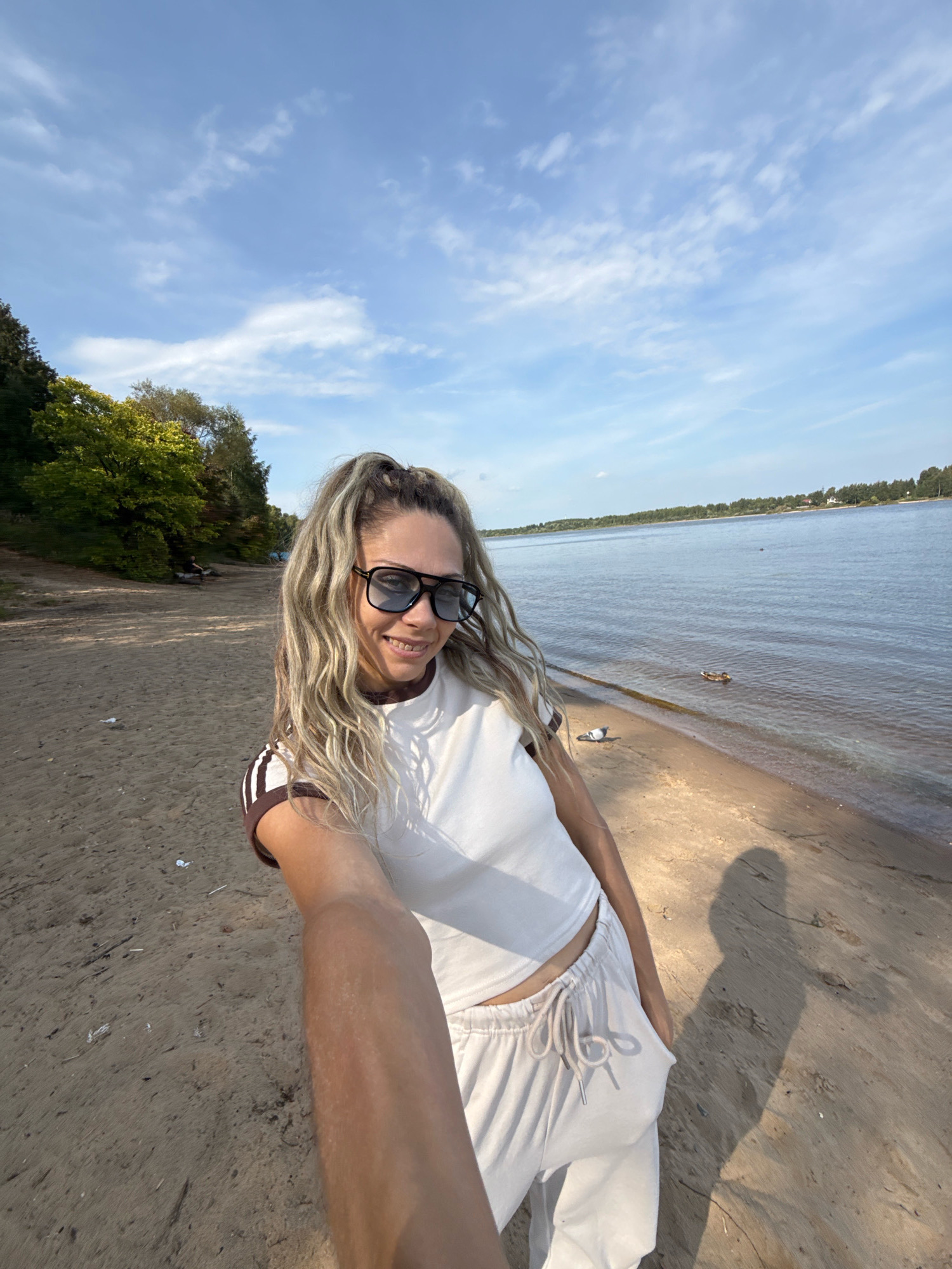 A woman with blonde, wavy hair tied in a high ponytail and wearing sunglasses, a white crop top with brown trim, and light-colored sweatpants takes a selfie on a sandy beach. The background shows a calm body of water, a tree-lined shore, and a blue sky with scattered clouds. In the water, a duck is visible. The scene appears to be in Serenovo, Russia.