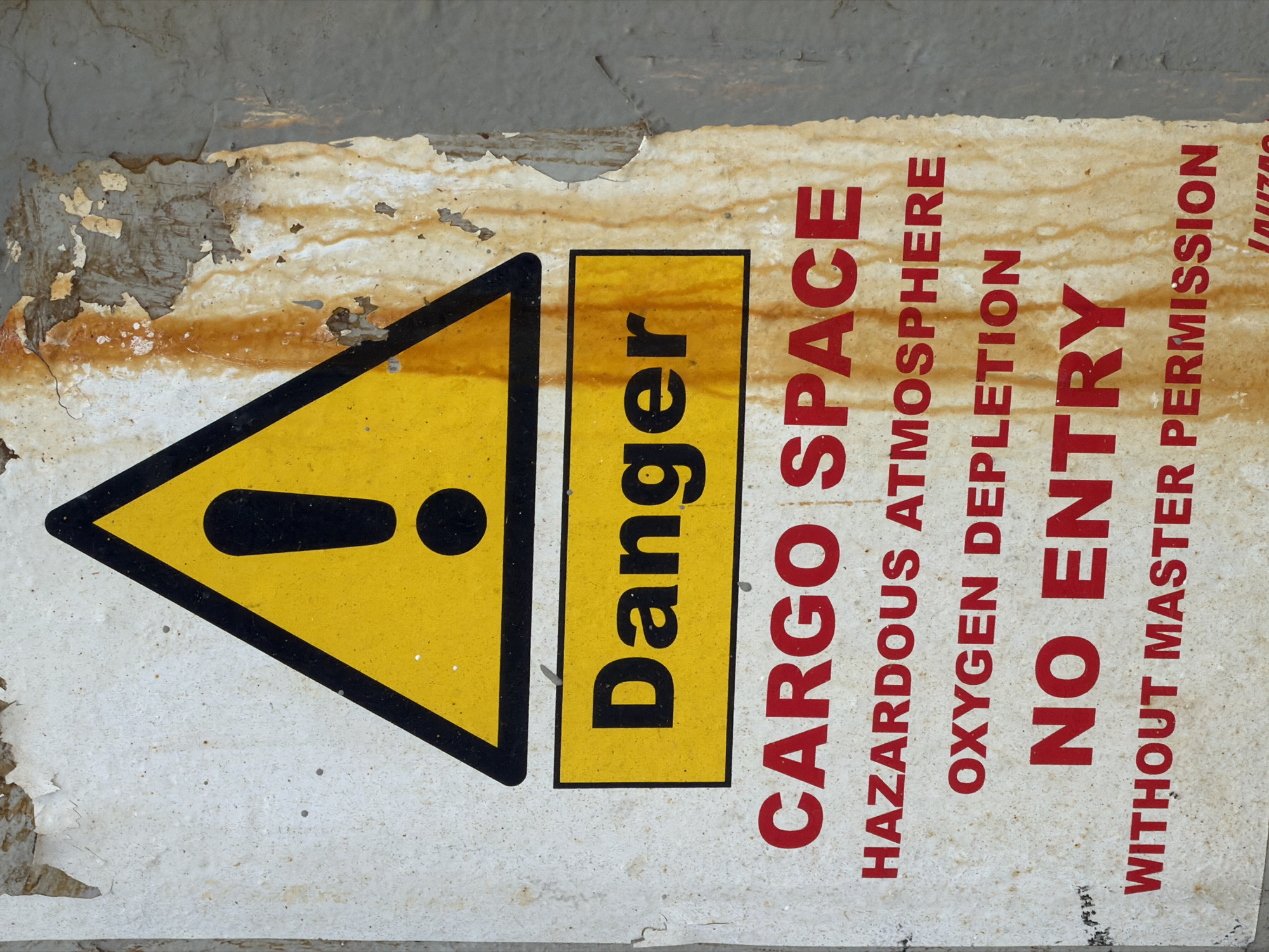 The image shows a weathered danger sign in Al Durra, Jordan. The sign is yellow with a black border and an exclamation mark inside a triangle, indicating a warning. Below the triangle, the word "Danger" is written in black on a yellow rectangle. The rest of the sign is white with red lettering. The text reads: "CARGO SPACE", "HAZARDOUS ATMOSPHERE", "OXYGEN DEPLETION", "NO ENTRY", and "WITHOUT MASTER PERMISSION". The sign is attached to a rough, textured wall that appears to be concrete or stone, with signs of rust and peeling paint. The overall condition of the sign suggests it has been exposed to the elements for a considerable time. There are no people or other objects visible in the immediate vicinity of the sign.